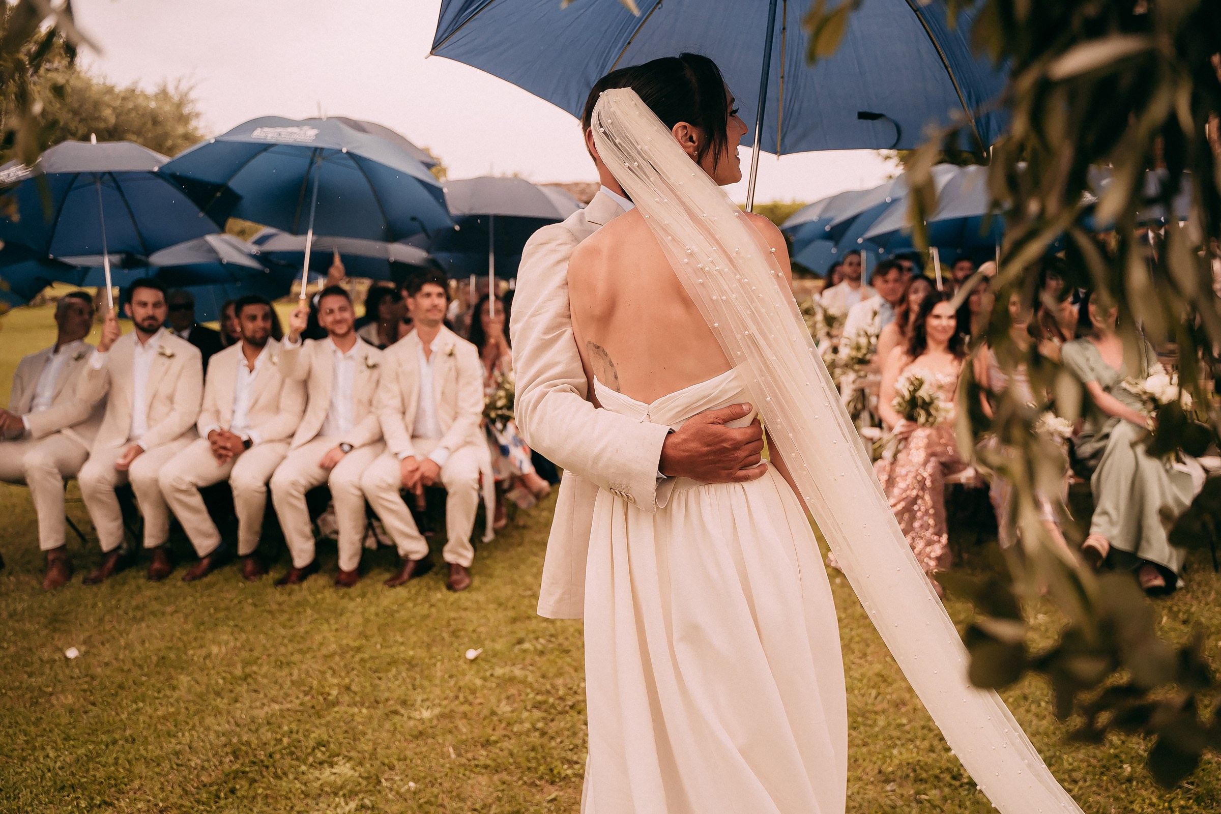 The bride and groom embrace under blue umbrellas, creating a romantic moment in the rain.