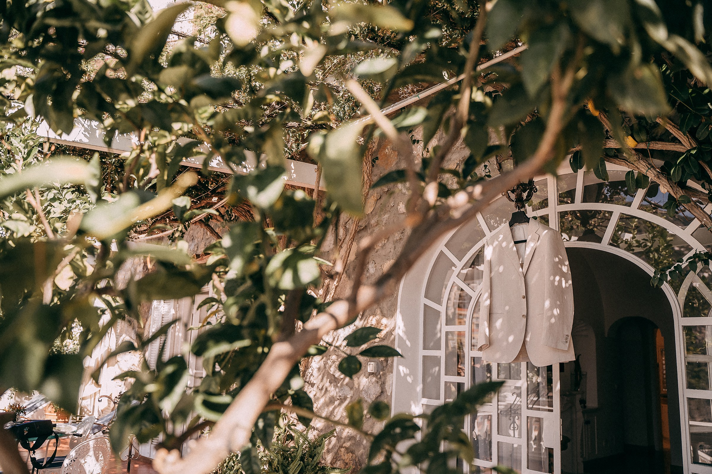 Two white suits hanging among the greenery at Villa Bohème, with an inviting glimpse of the villa’s arched doorway in the background.