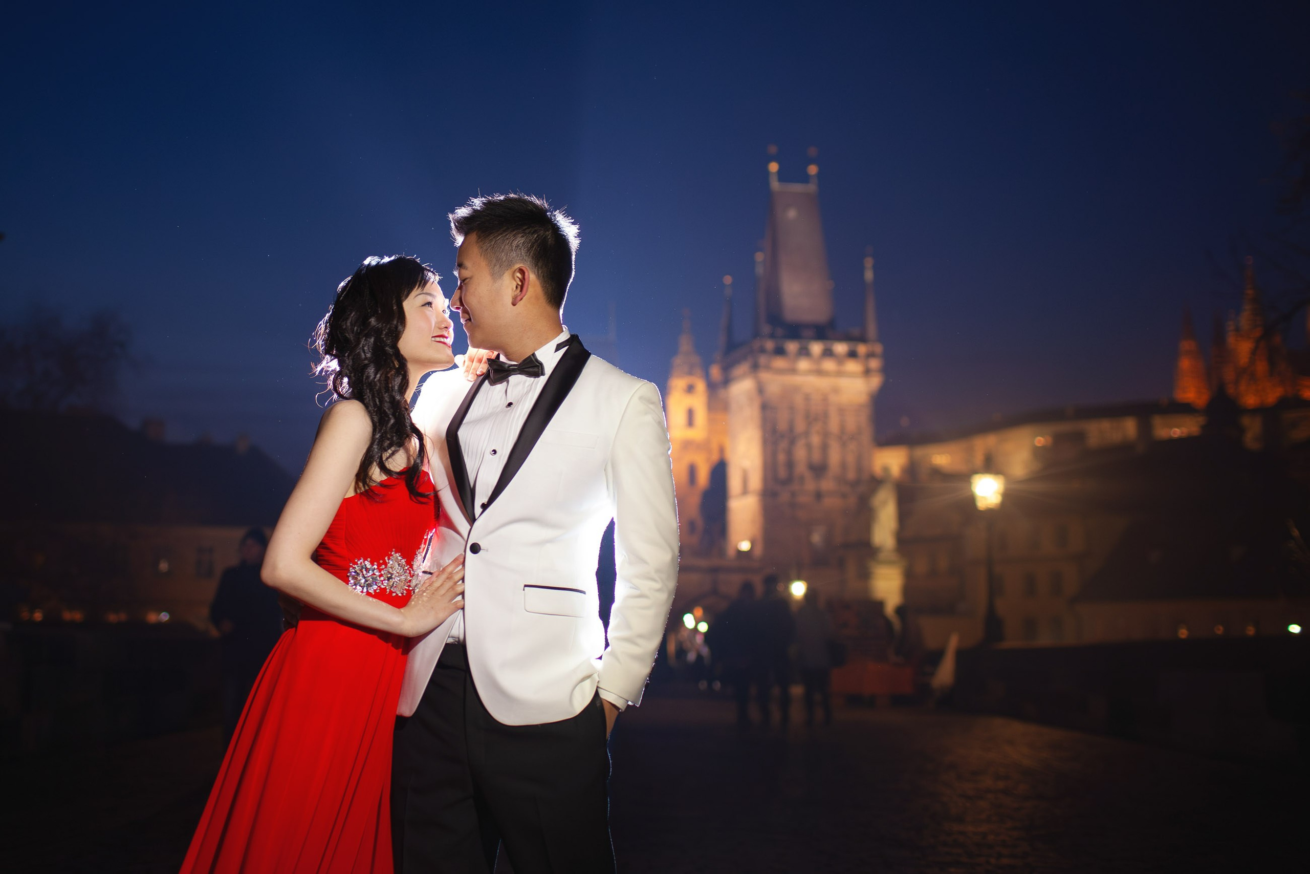 A smiling Hong Kong woman wearing a stylish red evening dress is held tight by her white tuxedo wearing man atop a misty Charles Bridge at night.