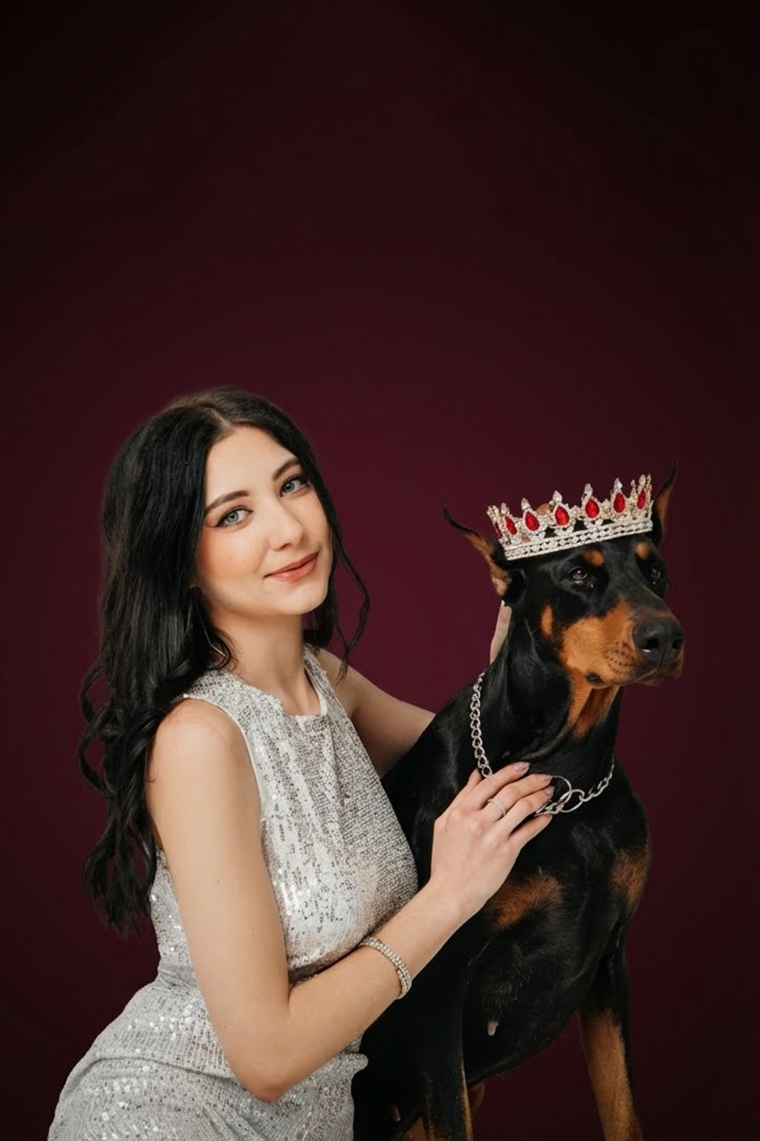 Frau im weißen Outfit sitzt neben einem Dobermann vor bordeauxrotem Hintergrund.