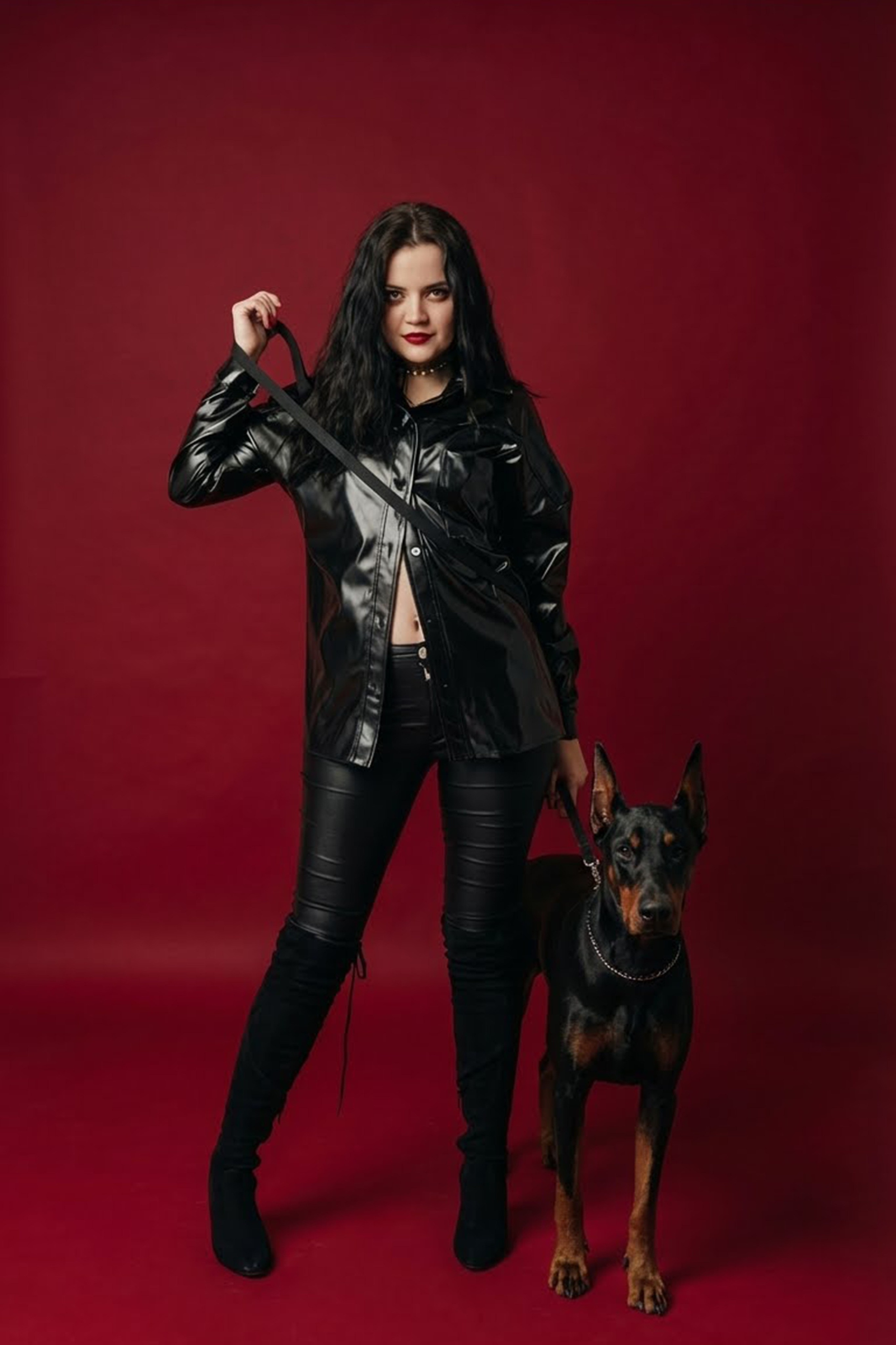 Frau in schwarzem Outfit steht mit einem Dobermann vor dunklem bordeauxrotem Hintergrund.