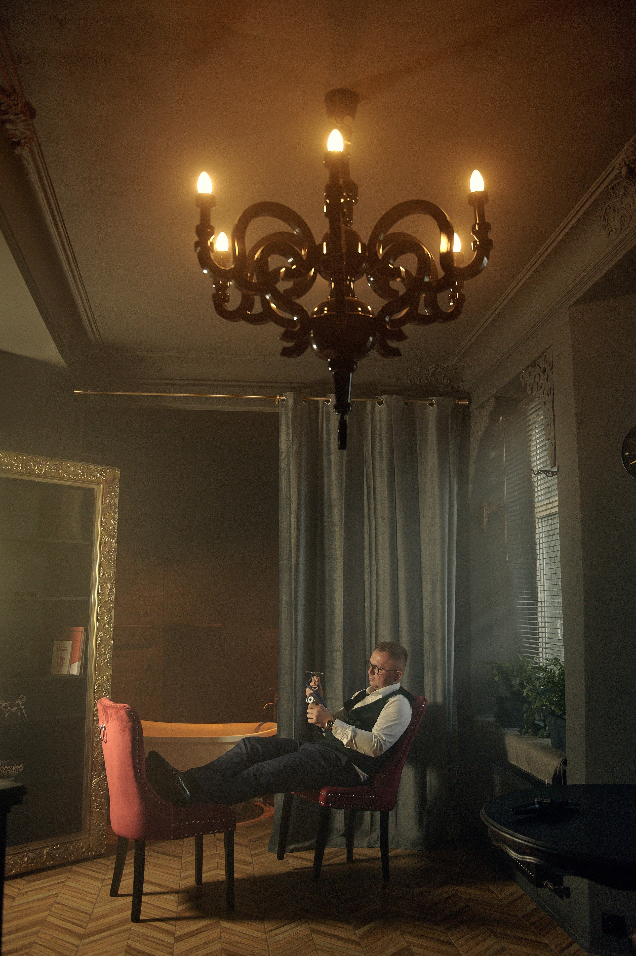 Stylizowany portret mężczyzny w okularach – siedzi z nogami na krześle, w tle lampka i złote lustro