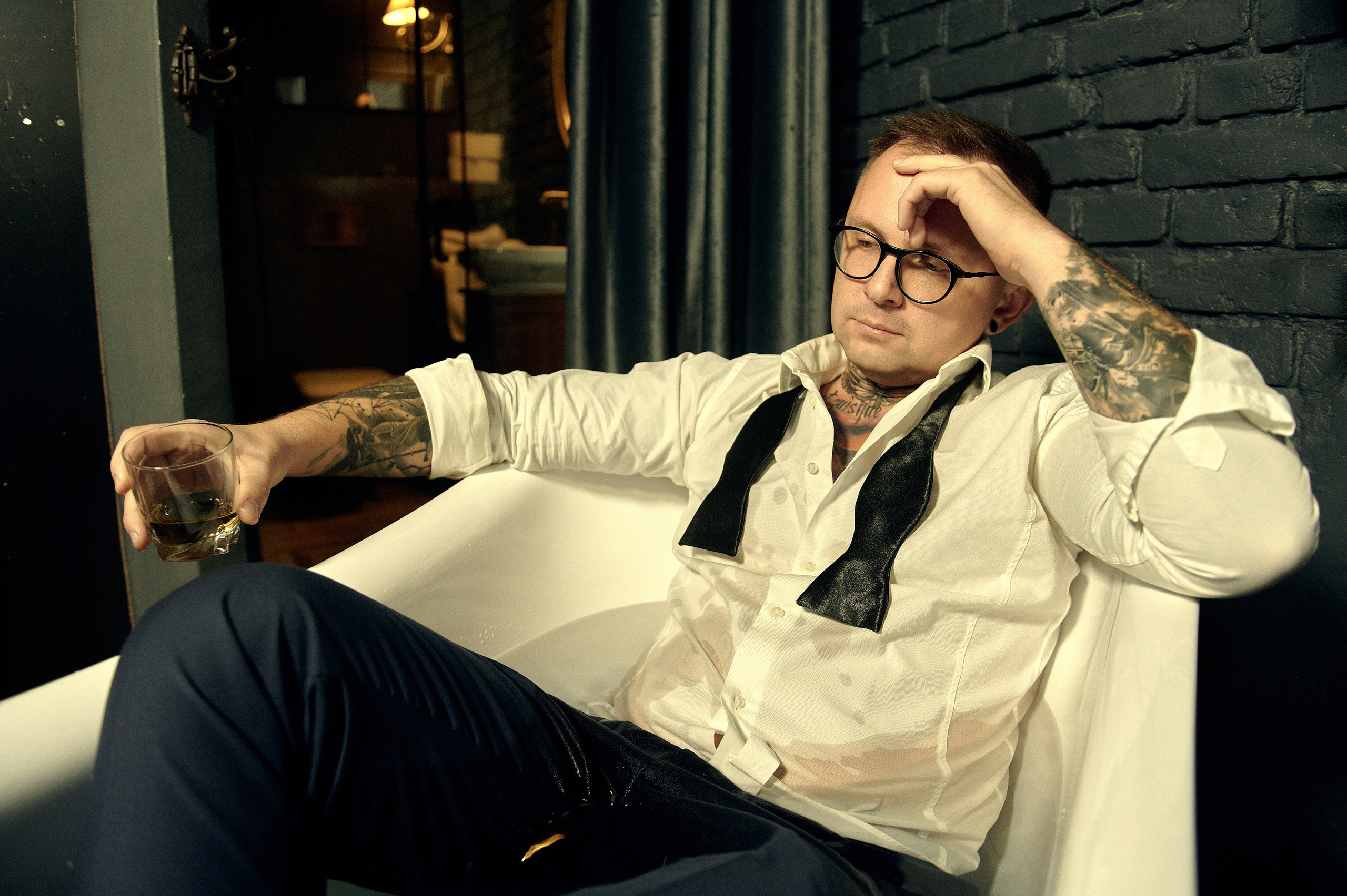 Mężczyzna w białej koszuli z rozwiązaną muchą siedzi w wannie, trzyma szklankę i wygląda na zamyślonego