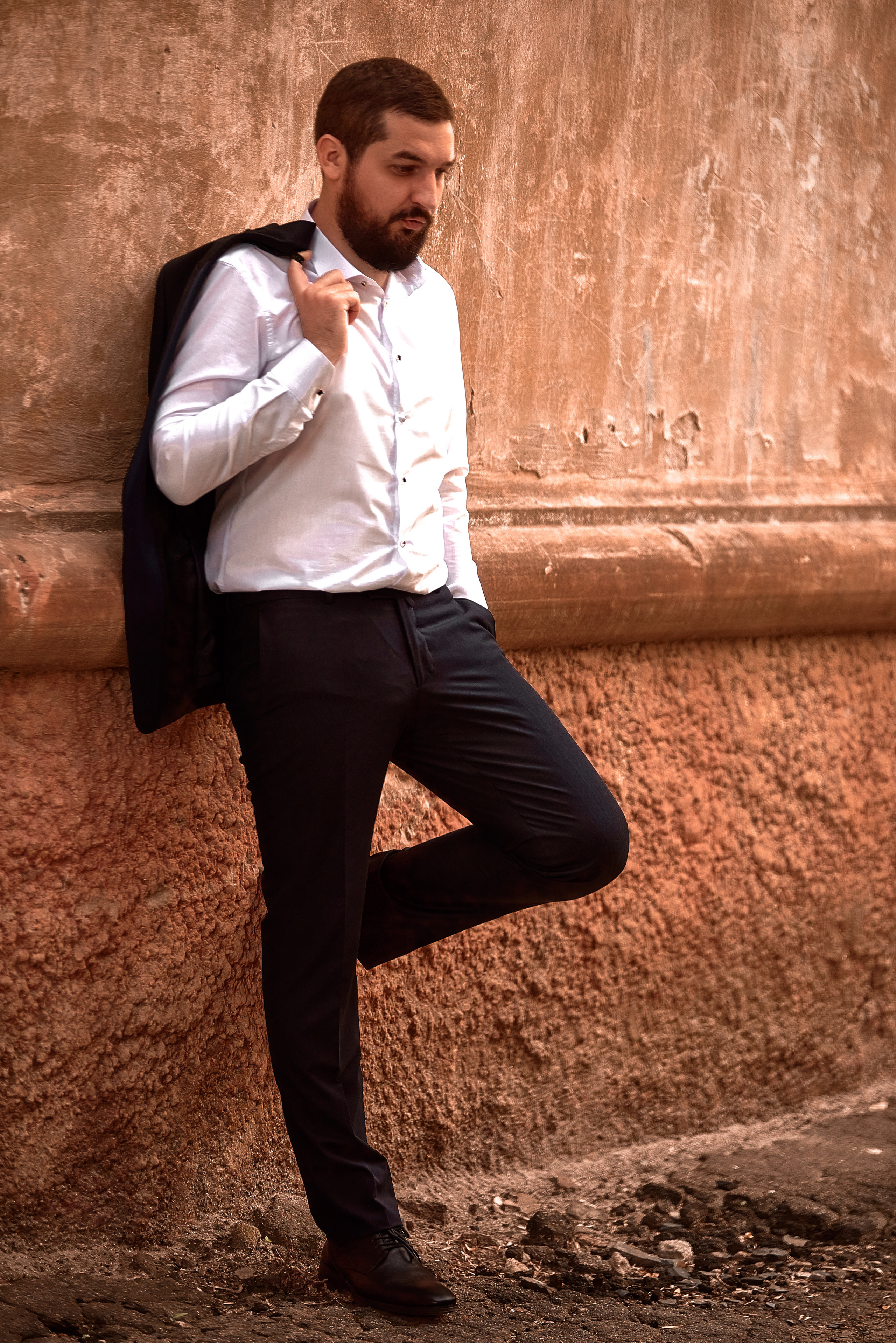 Мужской портрет в полный рост, парень с бородой в костюме с пиджаком на плече у винтажной стены.