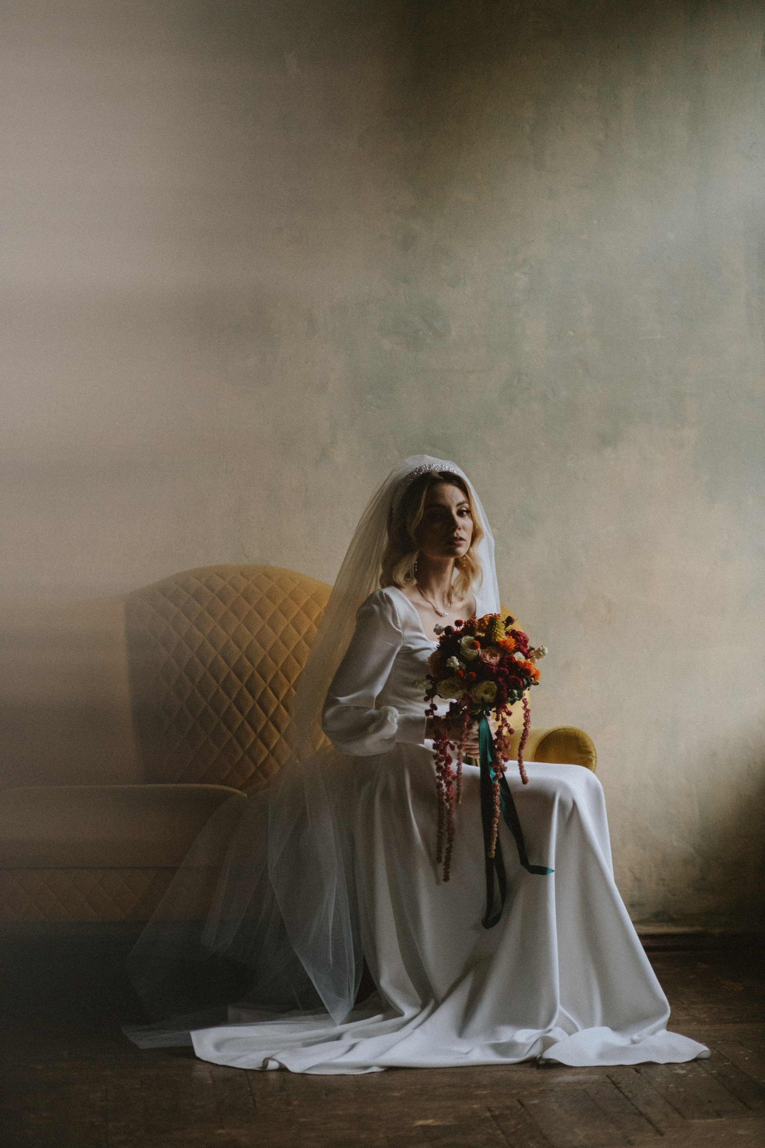 “Una sposa siede con grazia su una sedia gialla vintage, tenendo in mano un bouquet colorato con nastri lunghi e fluenti. Indossa un abito bianco a maniche lunghe, un diadema e un velo, guardando serenamente davanti a sé su uno sfondo morbido e neutro”.