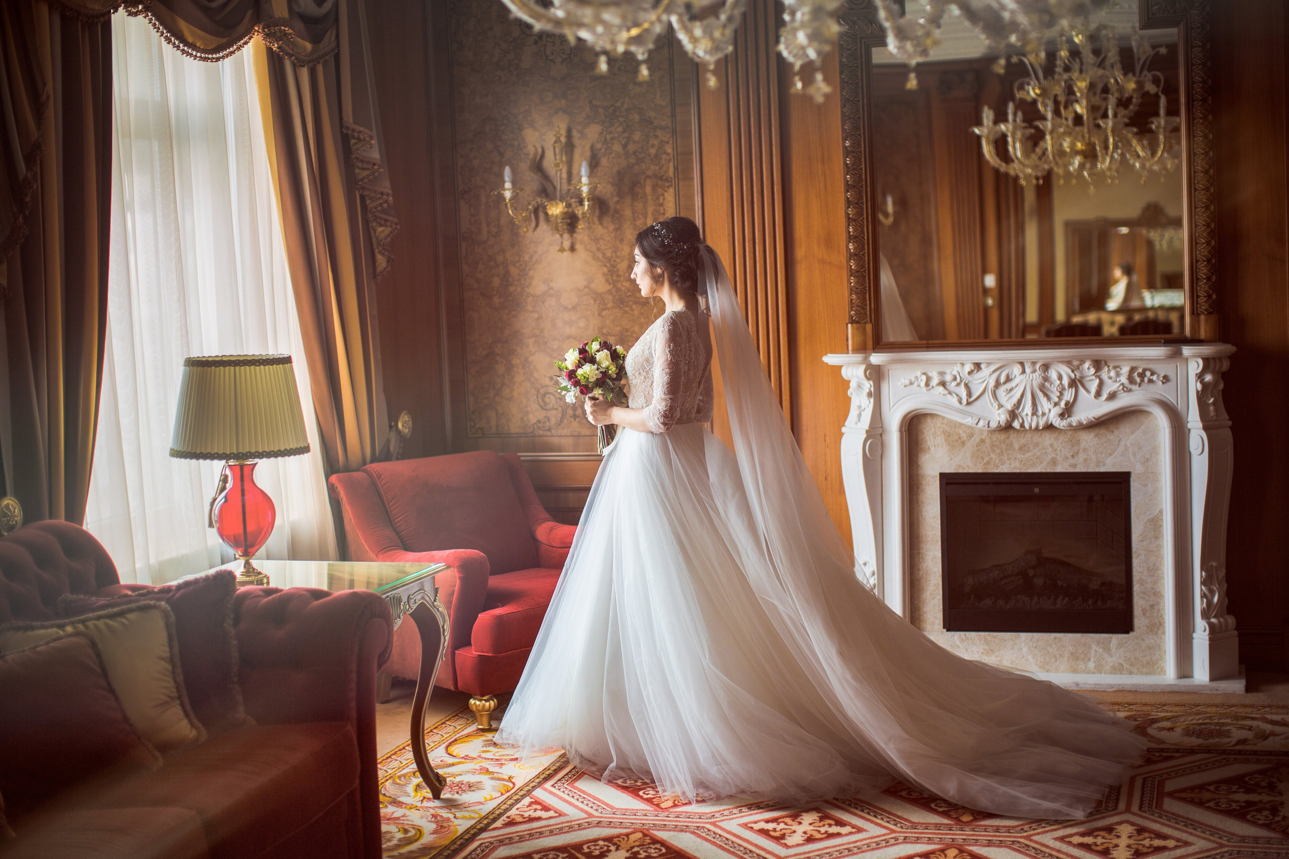 “Una bellissima sposa con un delicato top in pizzo e un fluente abito da sposa in tulle si affaccia con grazia alla finestra di una lussuosa stanza. Con in mano un bouquet di fiori vivaci, il suo velo ricade dietro di lei, aggiungendo eleganza a questo momento senza tempo, perfetto per un matrimonio romantico in Italia.”
