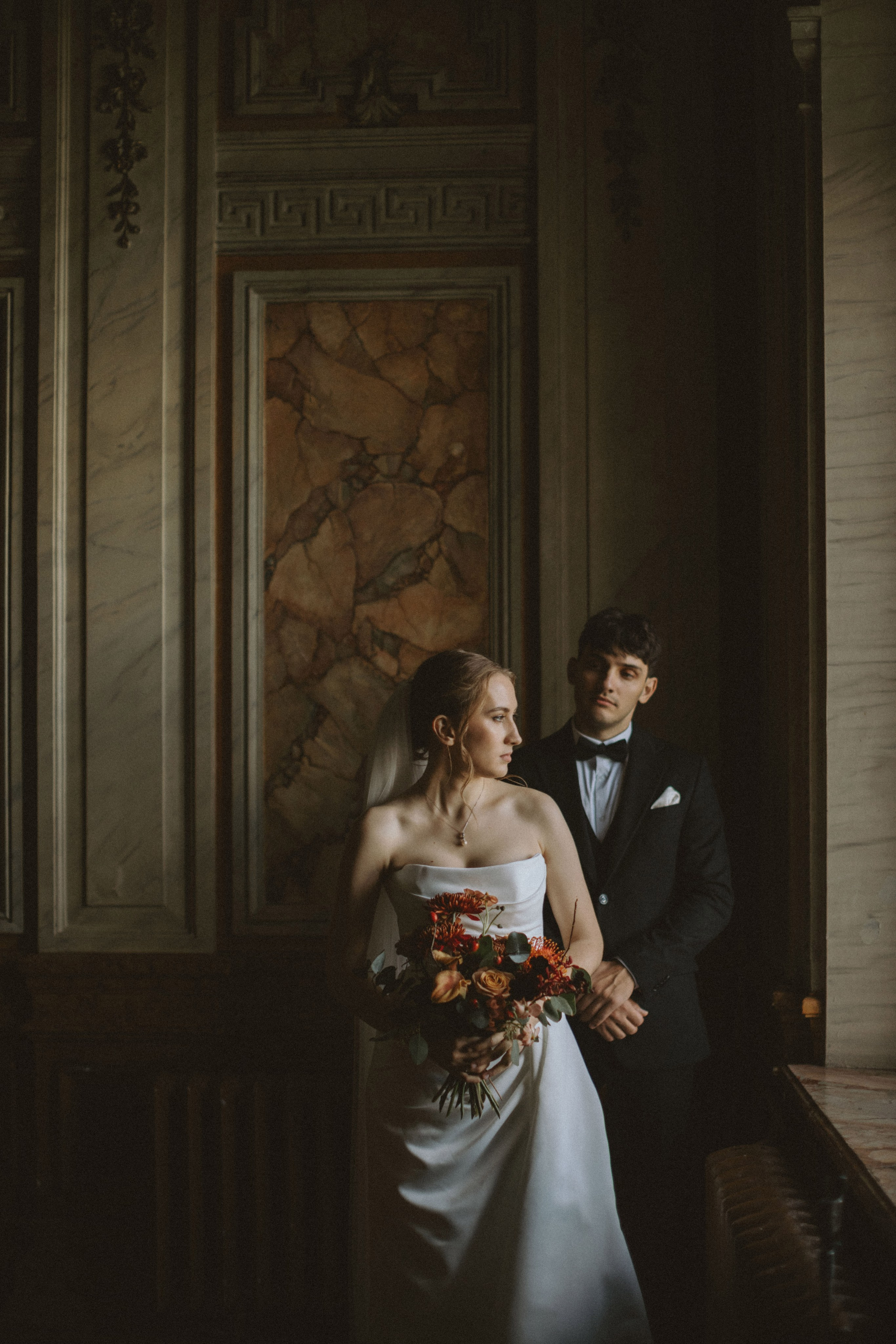 “Una coppia si trova in una stanza splendidamente illuminata, con lo sposo dietro la sposa. La sposa, che indossa un abito da sposa senza spalline, tiene in mano un vivace bouquet di fiori, mentre lo sposo, vestito in smoking, le sta vicino, guardandola con ammirazione. L'architettura elegante e classica della stanza, con le sue pareti di marmo e l'illuminazione calda, crea un'atmosfera romantica e intima per la scena”.