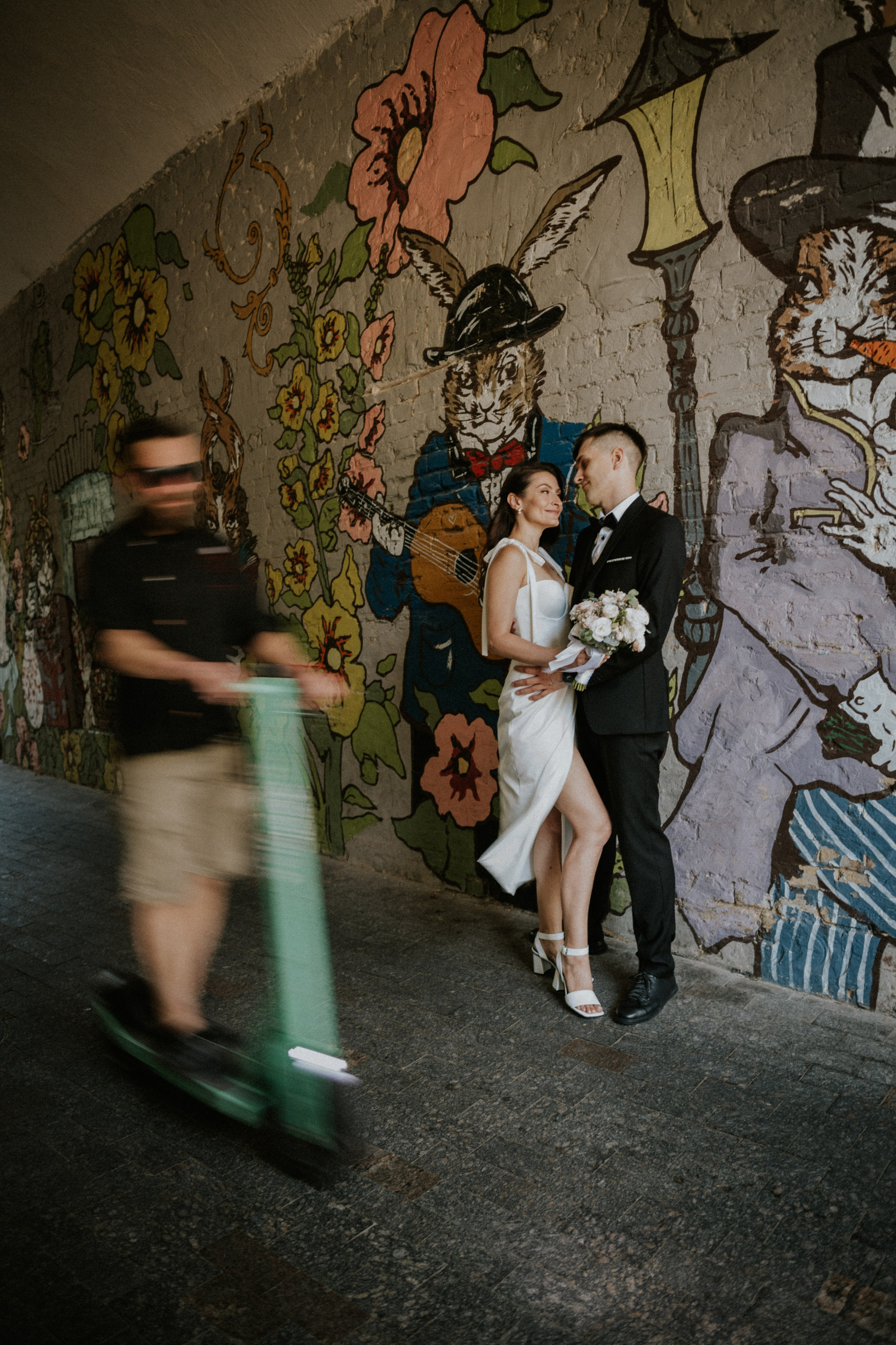 “Una sposa e uno sposo stanno vicini, sorridendo l'uno all'altro davanti a un murale colorato con conigli e fiori stravaganti. Un uomo su uno scooter elettrico passa sullo sfondo, creando un senso di movimento in questo momento vibrante e giocoso”.