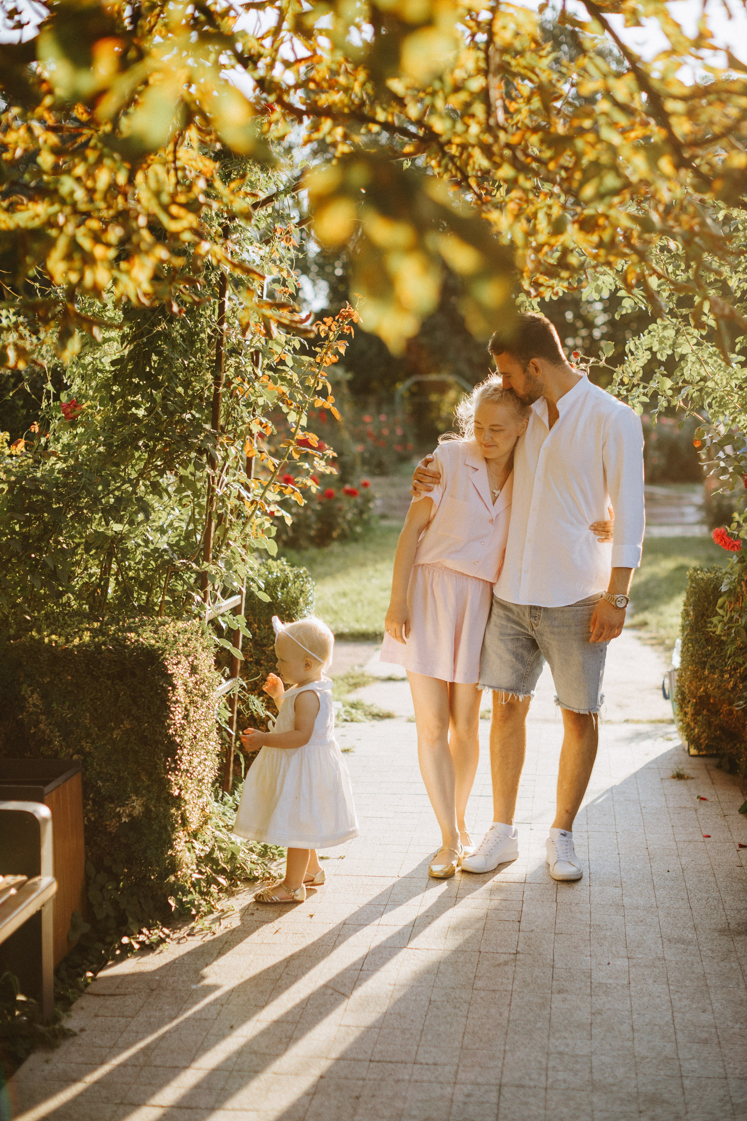 “Una famiglia gioiosa che si gode una giornata di sole all'aperto. Una bambina sorridente in abito bianco corre avanti mentre i genitori condividono un momento d'amore sullo sfondo, circondati dal verde e dalla luce del sole.”