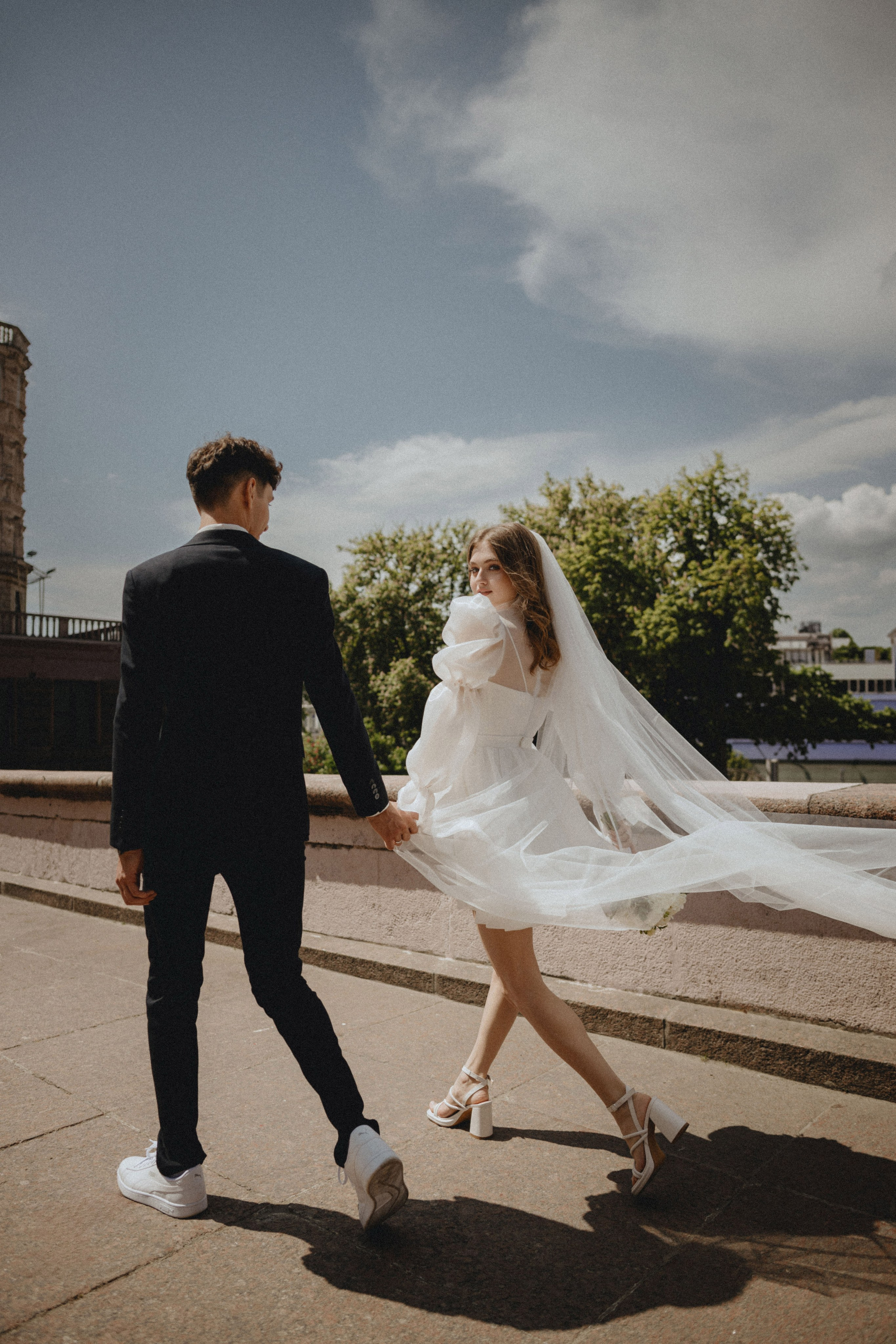 “Una sposa in abito bianco corto con maniche a sbuffo e scarpe da ginnastica cammina mano nella mano con lo sposo, che indossa un abito nero. Il velo della sposa scorre dietro di lei mentre gira la testa per sorridere a lui. Camminano lungo un sentiero soleggiato con alberi e un cielo luminoso sullo sfondo”.