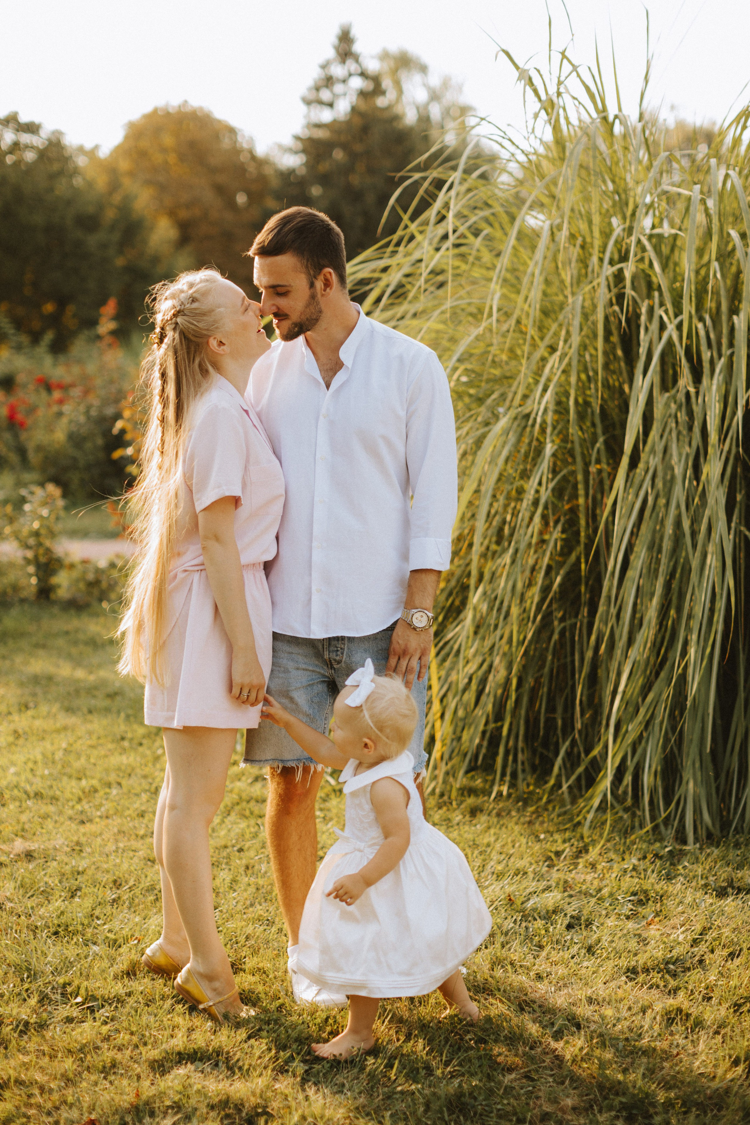 “Un gioioso momento di famiglia catturato all'aperto con la luce soffusa del sole. Una coppia è vicina, sorridendo l'uno all'altra con la figlia che li tiene per mano. La donna, vestita con un abito rosa, guarda amorevolmente il suo partner, mentre l'uomo, in camicia bianca e pantaloncini, la guarda con affetto. La loro giovane figlia, vestita con un abito bianco e con un fiocco in testa, sta a piedi nudi sull'erba e tende la mano alla madre. Lo sfondo è caratterizzato da un fogliame verde e rigoglioso, che crea un'atmosfera calda e intima”.