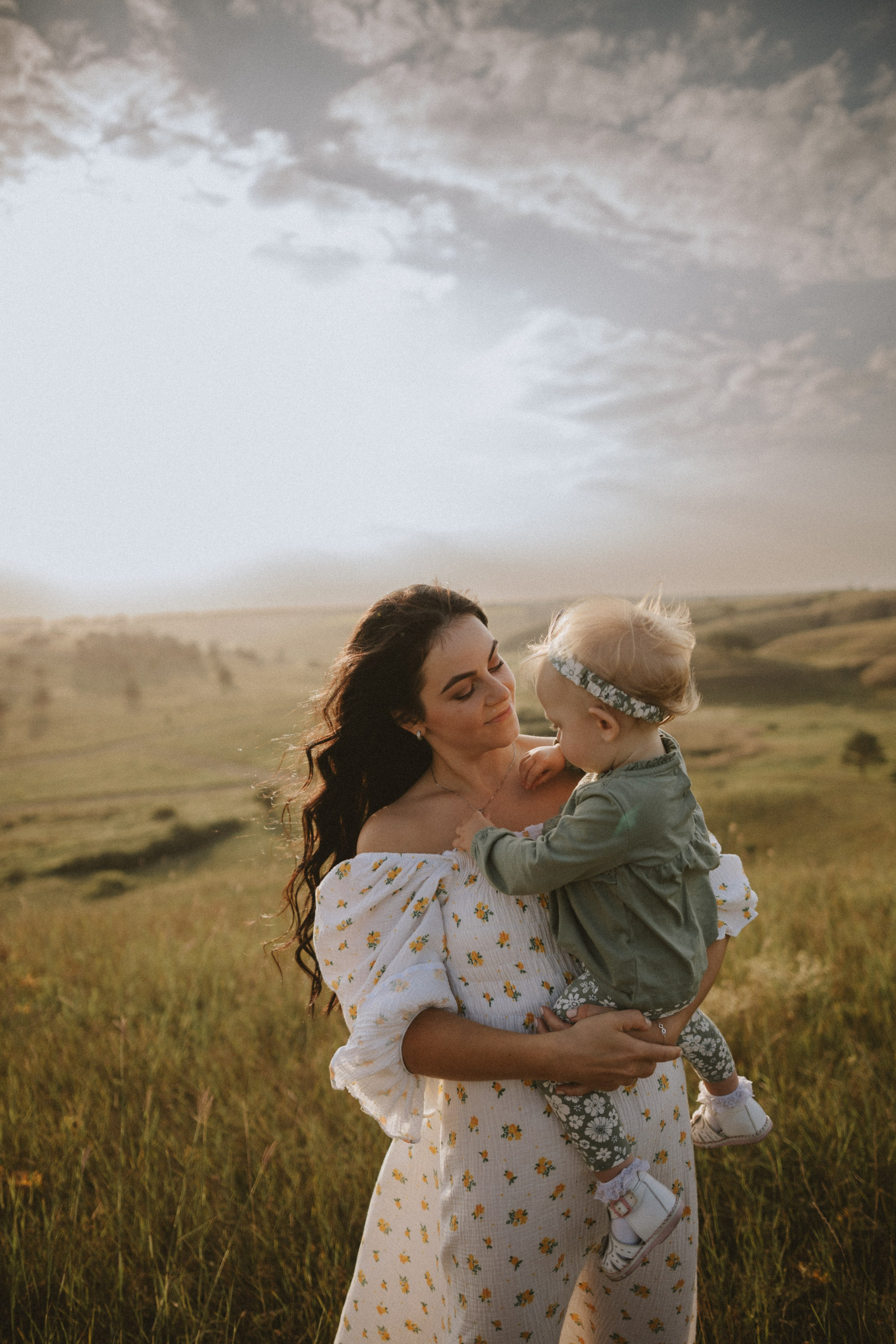 “Una madre che tiene in braccio il suo bambino su una splendida collina erbosa, con il bambino che indossa un maglione verde e leggings floreali. Lo sfondo è caratterizzato da un tramonto dorato con colline ondulate, e la madre guarda amorevolmente il suo bambino mentre lo tiene delicatamente in braccio.”