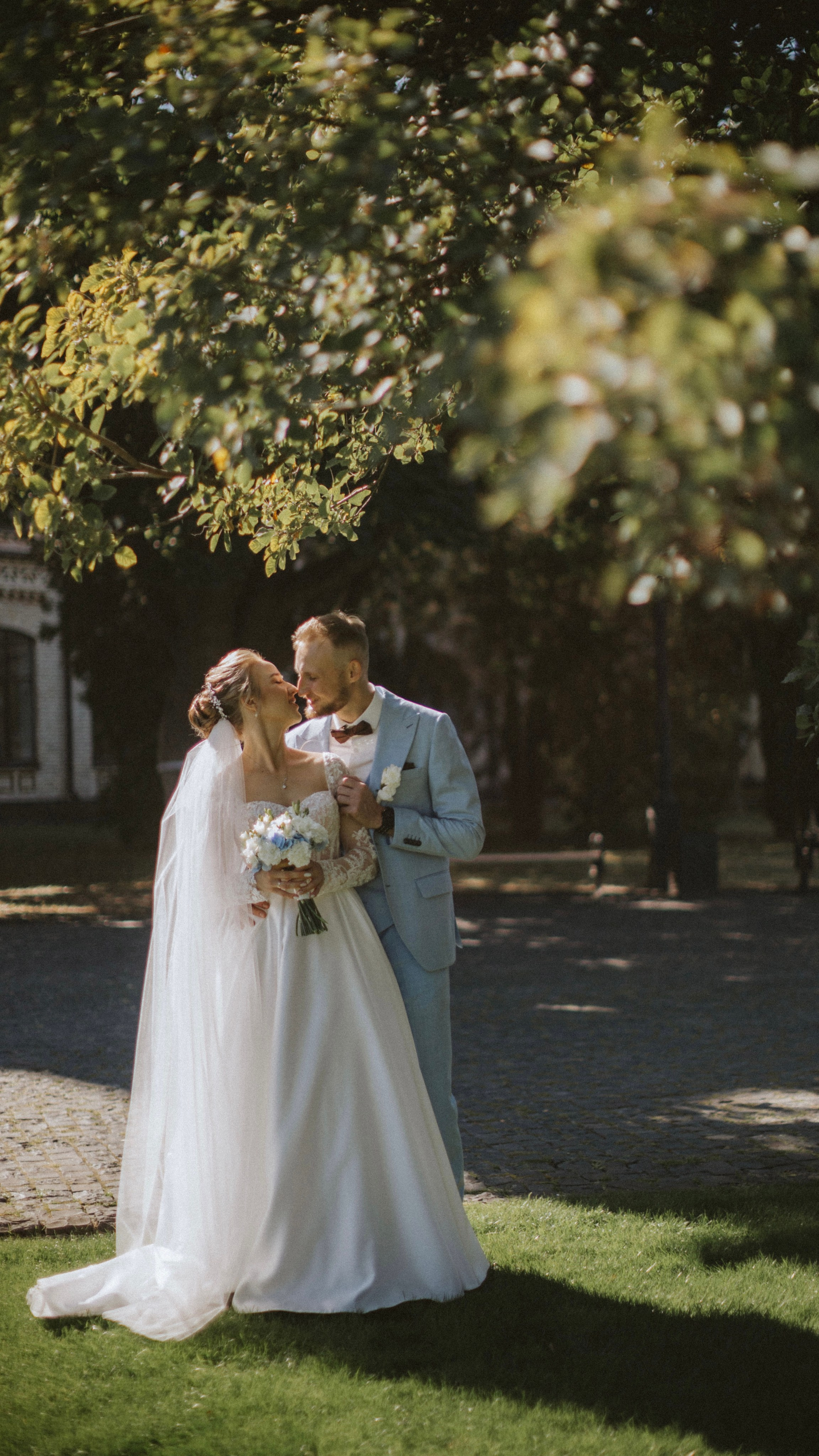 “Una sposa e uno sposo si scambiano un tenero bacio sotto un albero, circondati dalla luce del sole e dal verde. La sposa, in un classico abito bianco con dettagli in pizzo, tiene in mano un bouquet di fiori bianchi e blu. Lo sposo, vestito con un abito azzurro con papillon, la abbraccia dolcemente. La morbida luce del sole filtra attraverso le foglie, creando un'atmosfera romantica e serena”.