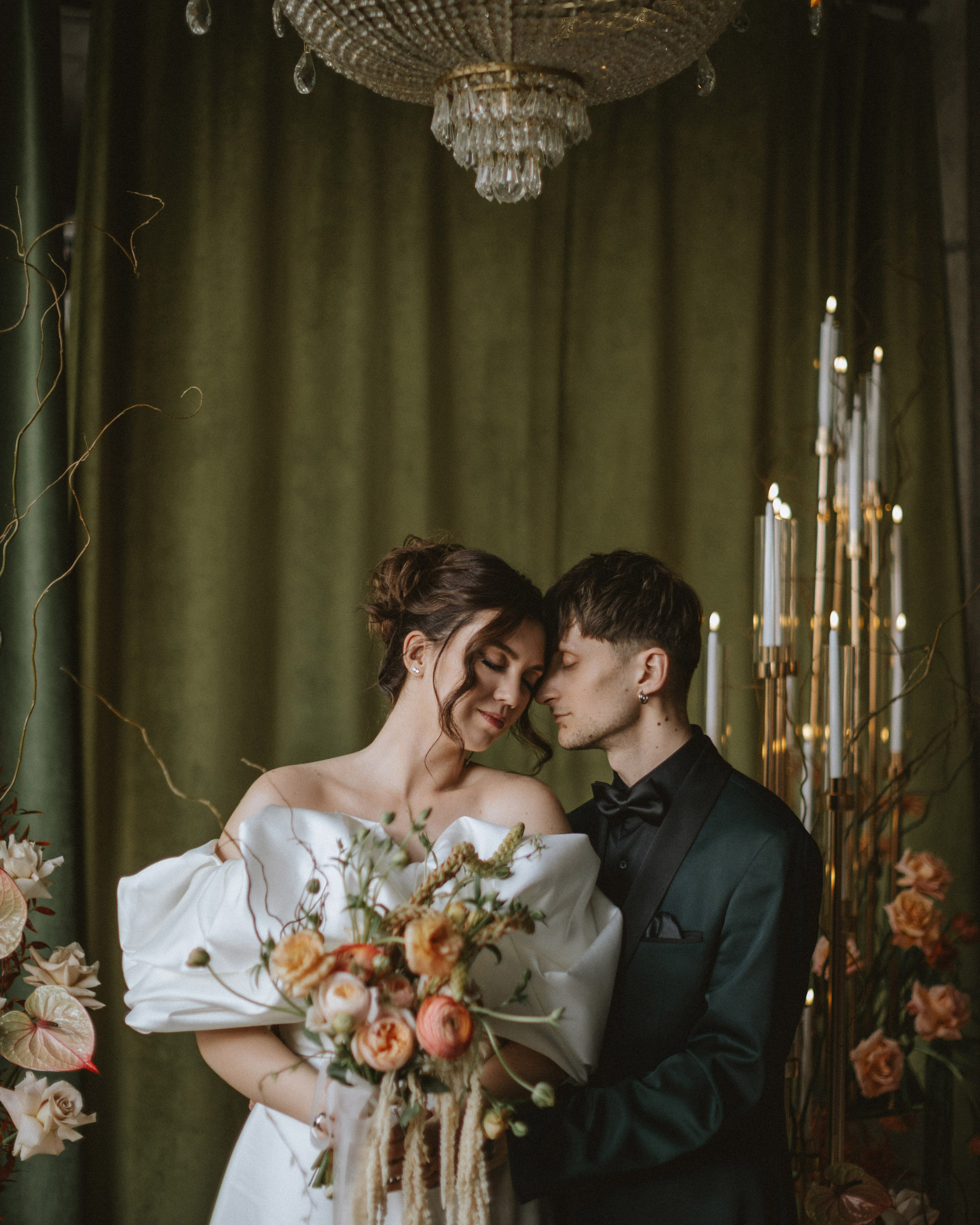 “The bride and groom in elegant attire embrace, the bride holding a bouquet with pink and orange flowers against the backdrop of green curtains and a chandelier.”
