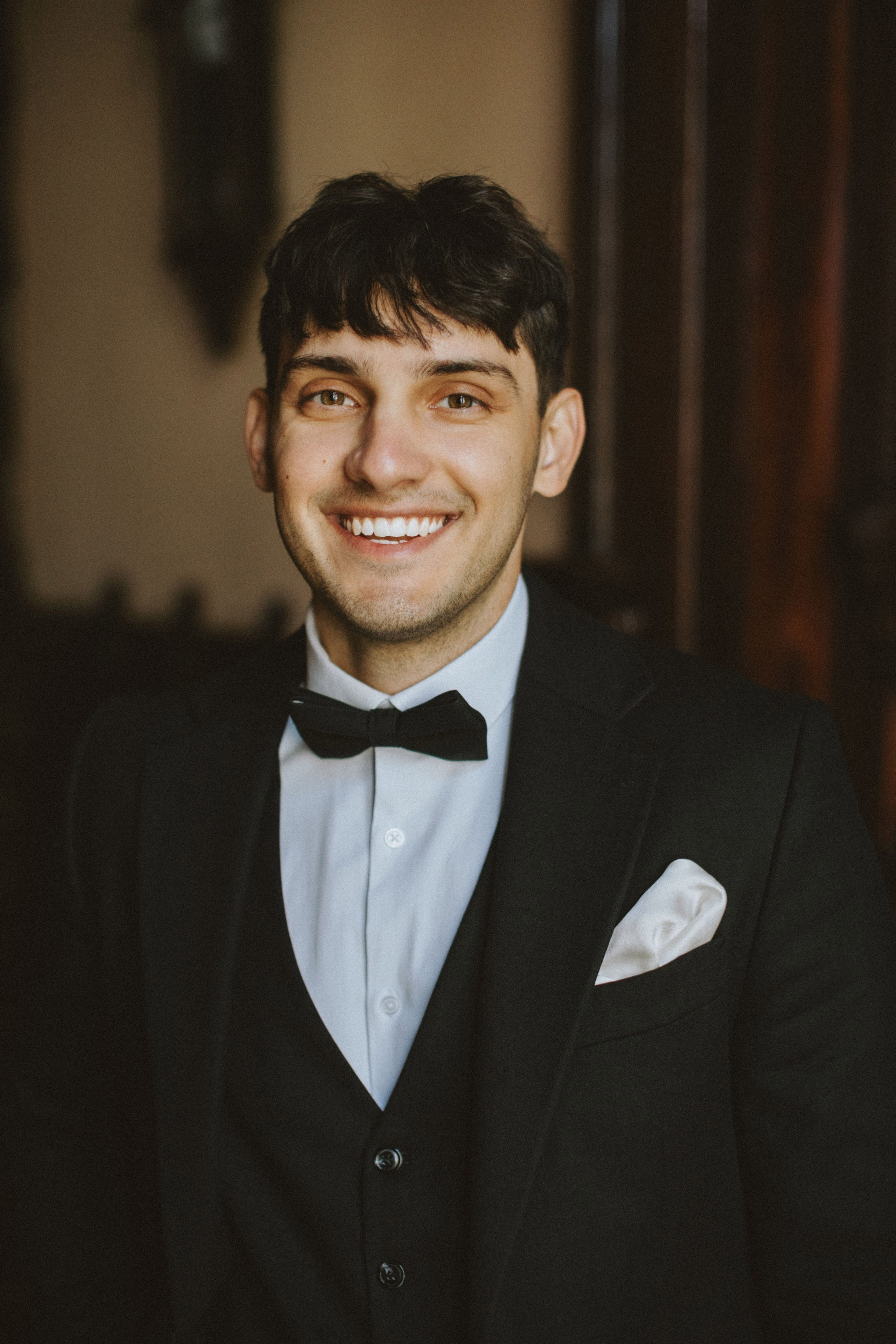 “Primo piano di un uomo sorridente in smoking, con papillon e fazzoletto da taschino bianco. I suoi capelli sono corti e acconciati con cura. Appare gioioso e sicuro di sé, con un sorriso smagliante, in piedi in una stanza ben illuminata con dettagli interni classici sullo sfondo”.