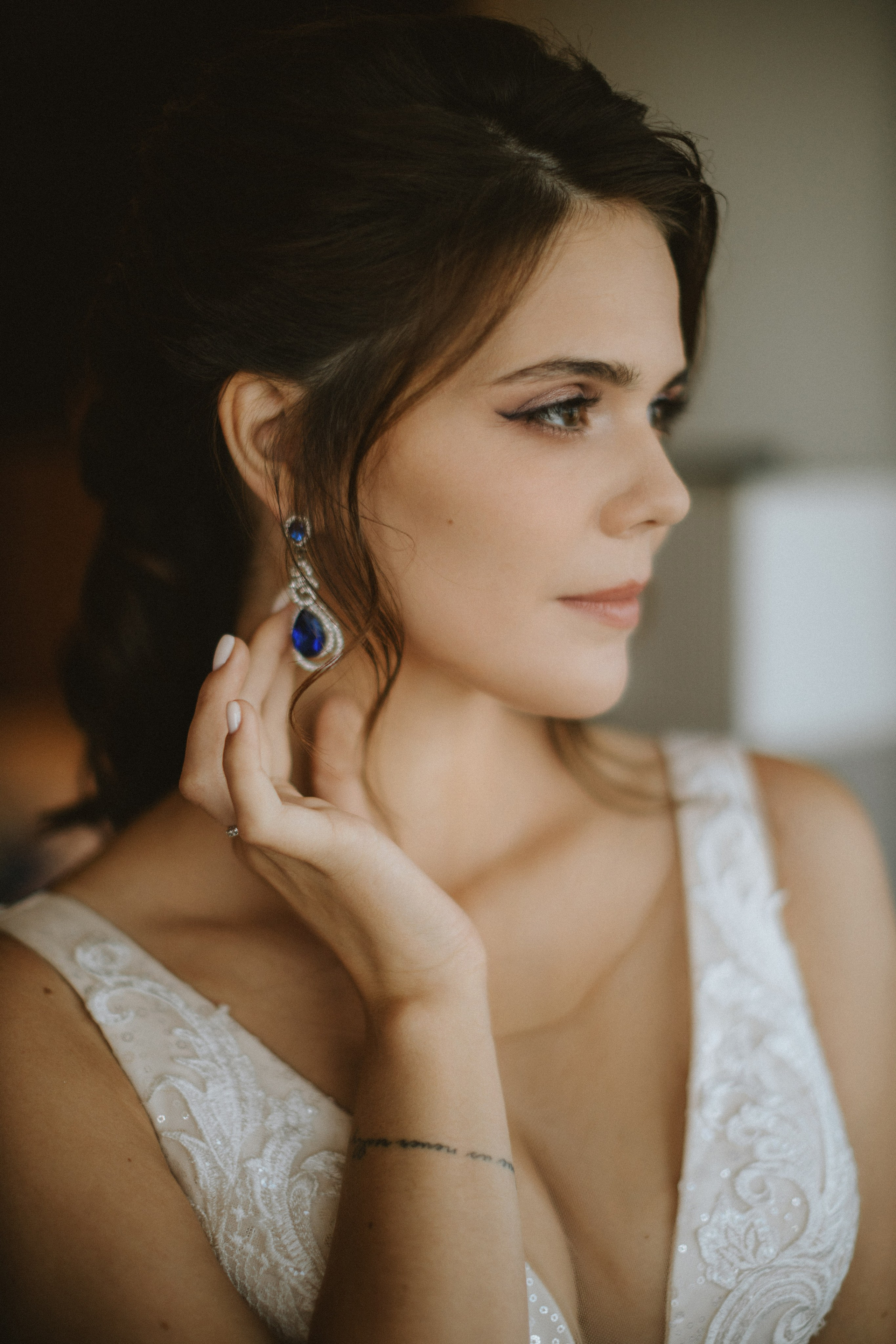 “Primo piano di una sposa che si aggiusta delicatamente l'orecchino con una splendida gemma blu. Indossa un abito da sposa in pizzo e i suoi capelli sono acconciati in un'elegante pettinatura. La luce soffusa mette in risalto i suoi lineamenti aggraziati e gli intricati dettagli dell'abito da sposa”.