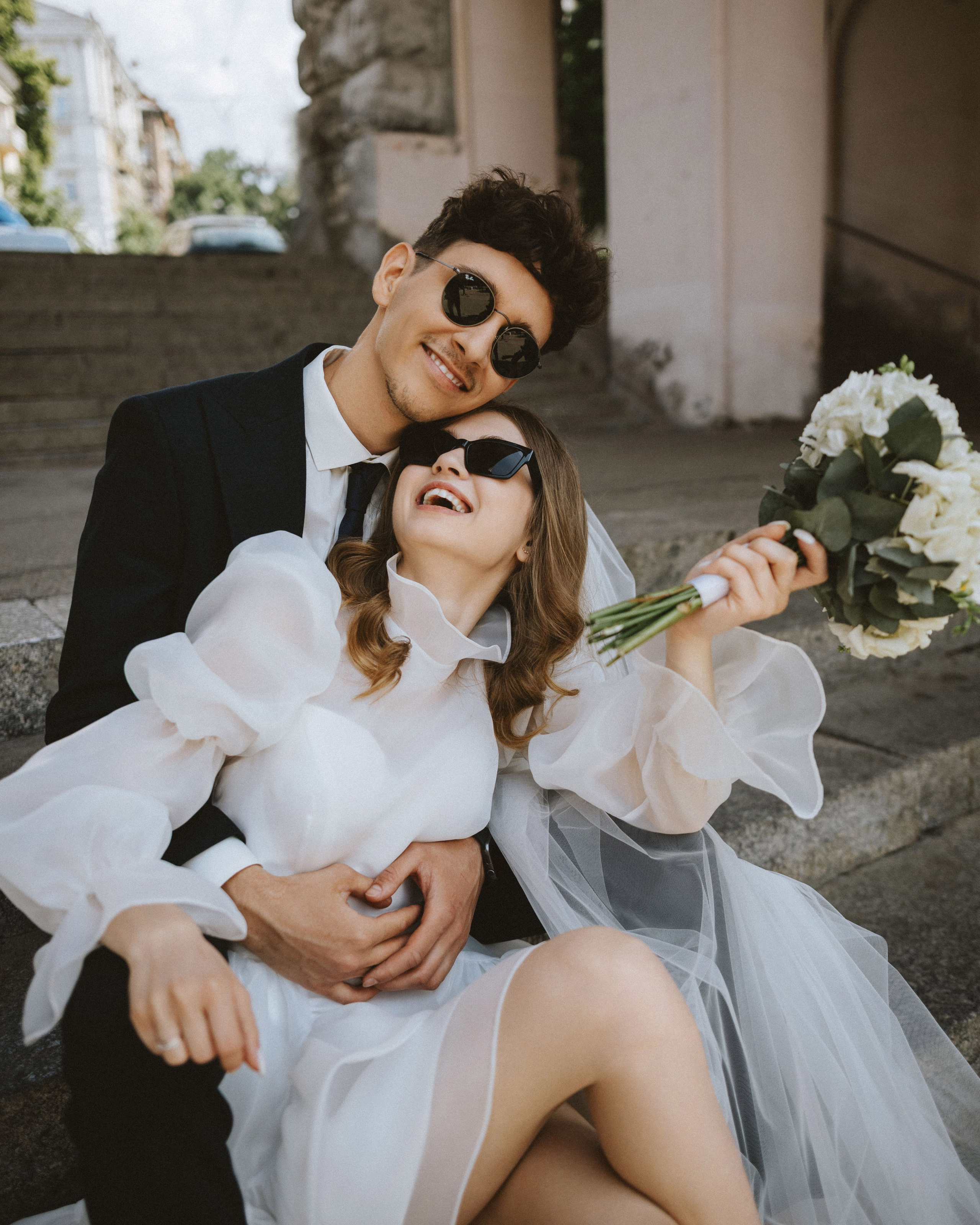 “Una coppia si gode un momento di gioco all'aperto, seduta su una serie di scale in pietra. La sposa, che indossa un elegante abito da sposa bianco con maniche a sbuffo e occhiali da sole, ride gioiosamente tenendo in mano un bouquet di fiori bianchi. Il suo compagno, vestito con un completo e occhiali da sole, sorride ampiamente mentre la abbraccia, creando un'atmosfera divertente e spensierata”.