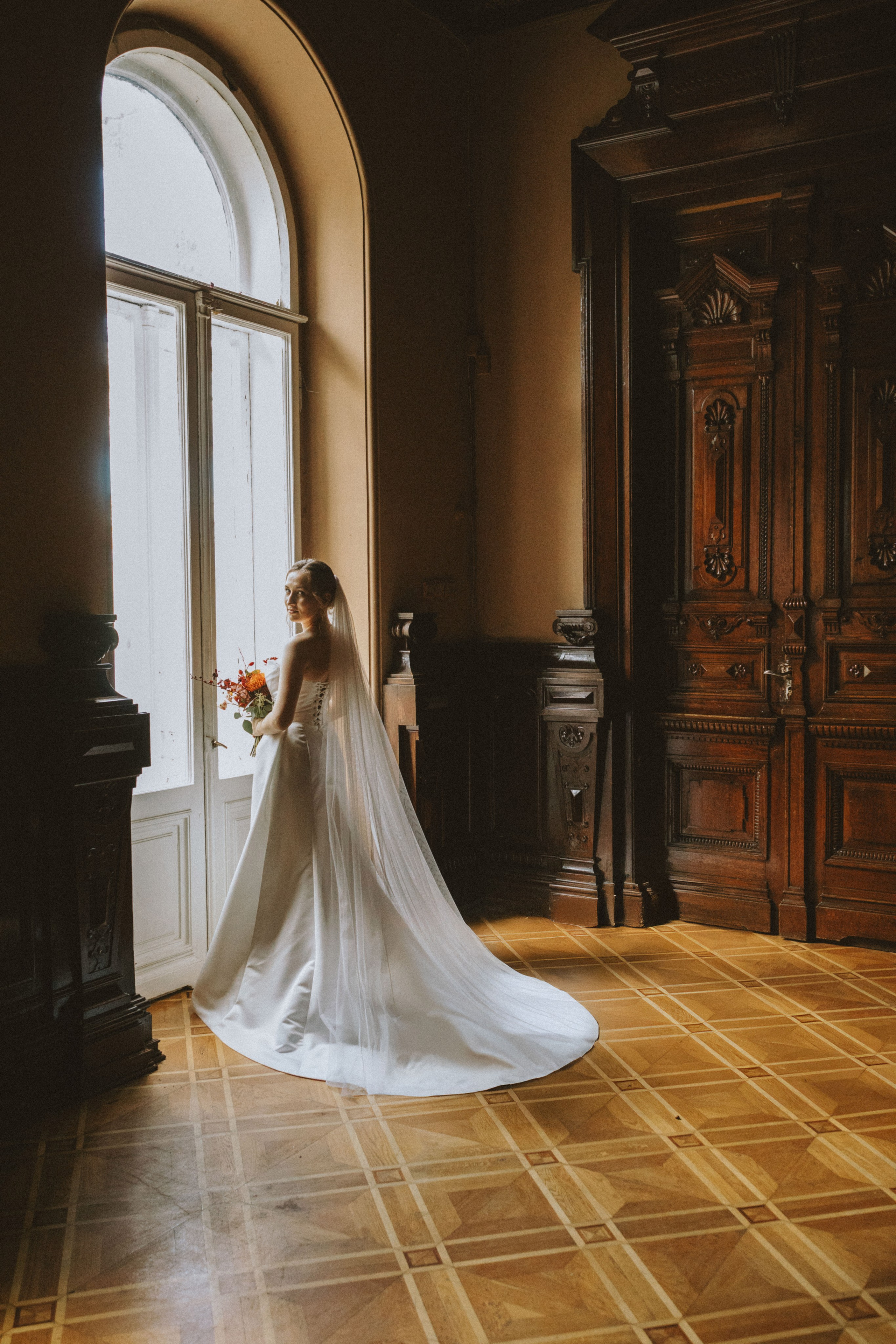 “Una sposa in un elegante abito bianco si trova vicino a una grande finestra in una stanza elegante e vintage. Guarda fuori dalla finestra, tenendo in mano un bouquet di fiori, mentre il lungo velo le scende lungo la schiena. L'arredamento in legno e il pavimento intricato della stanza creano un'atmosfera calda e romantica”.