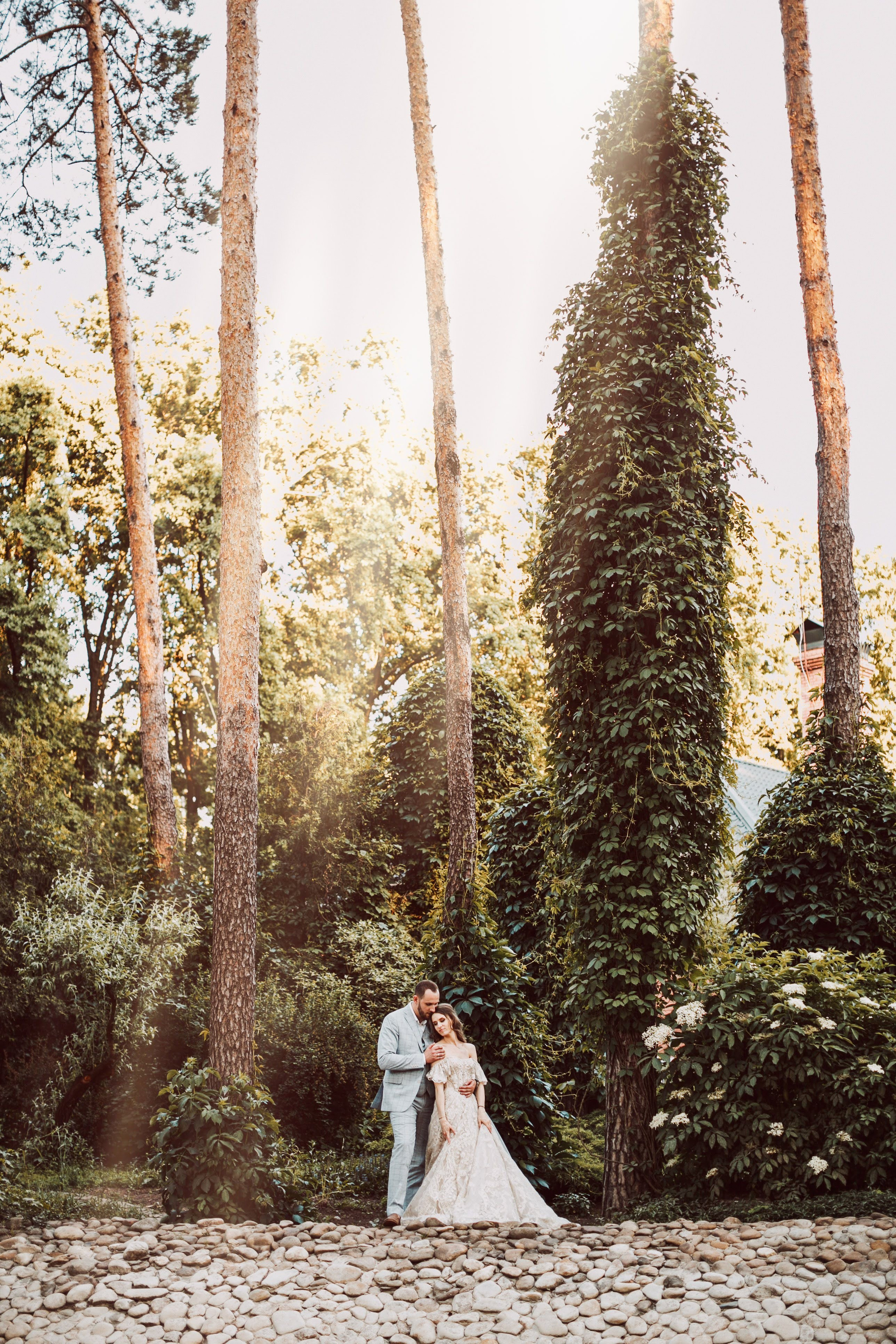 “Una coppia si trova insieme in una foresta serena, circondata da alti alberi e da una vegetazione lussureggiante. La sposa, che indossa un bellissimo abito di pizzo, si appoggia delicatamente allo sposo, vestito con un abito grigio chiaro. La luce del sole filtra attraverso gli alberi, proiettando una luce calda sulla coppia, creando un'atmosfera romantica e pacifica. Il terreno sotto di loro è ricoperto di pietre lisce, che aggiungono un tocco naturale alla scena”.