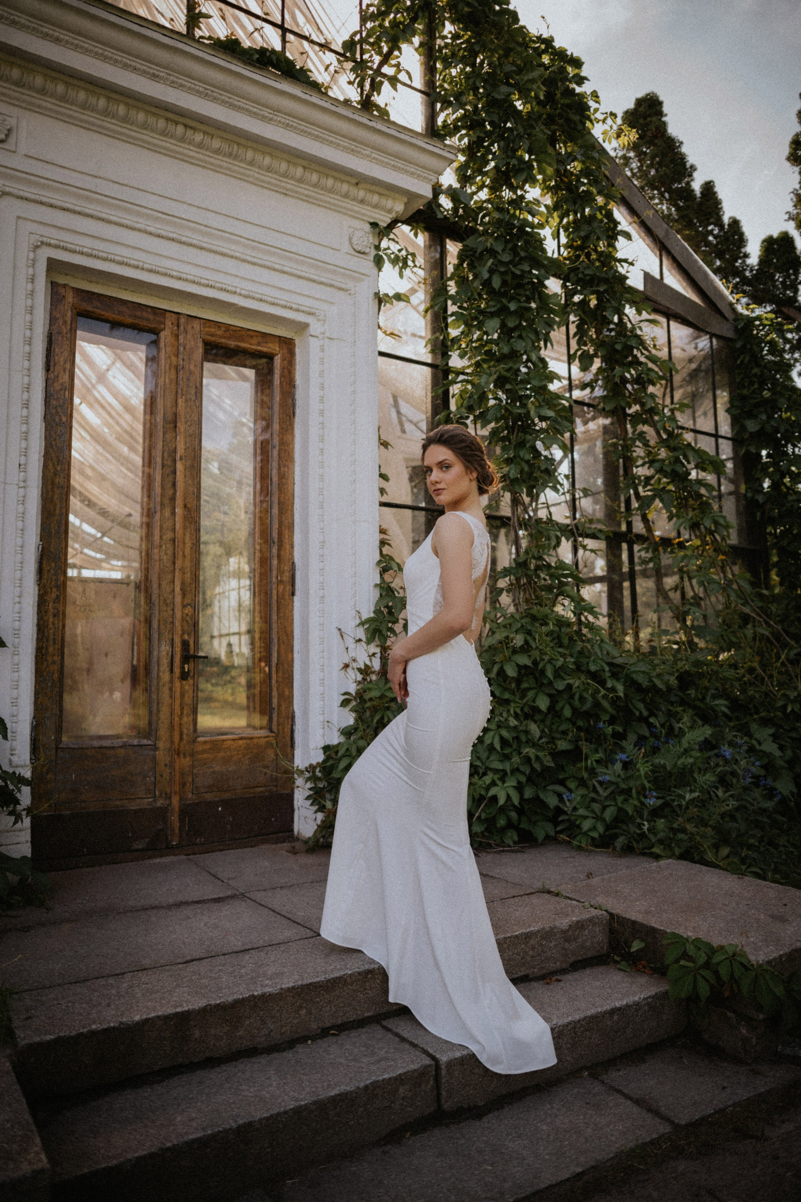 “Una sposa in piedi con grazia sui gradini di un bellissimo edificio ricoperto di edera. Indossa un abito bianco semplice e aderente che mette in risalto i delicati dettagli in pizzo sulla schiena. La luce naturale filtra attraverso il verde, creando un'atmosfera serena ed elegante”.