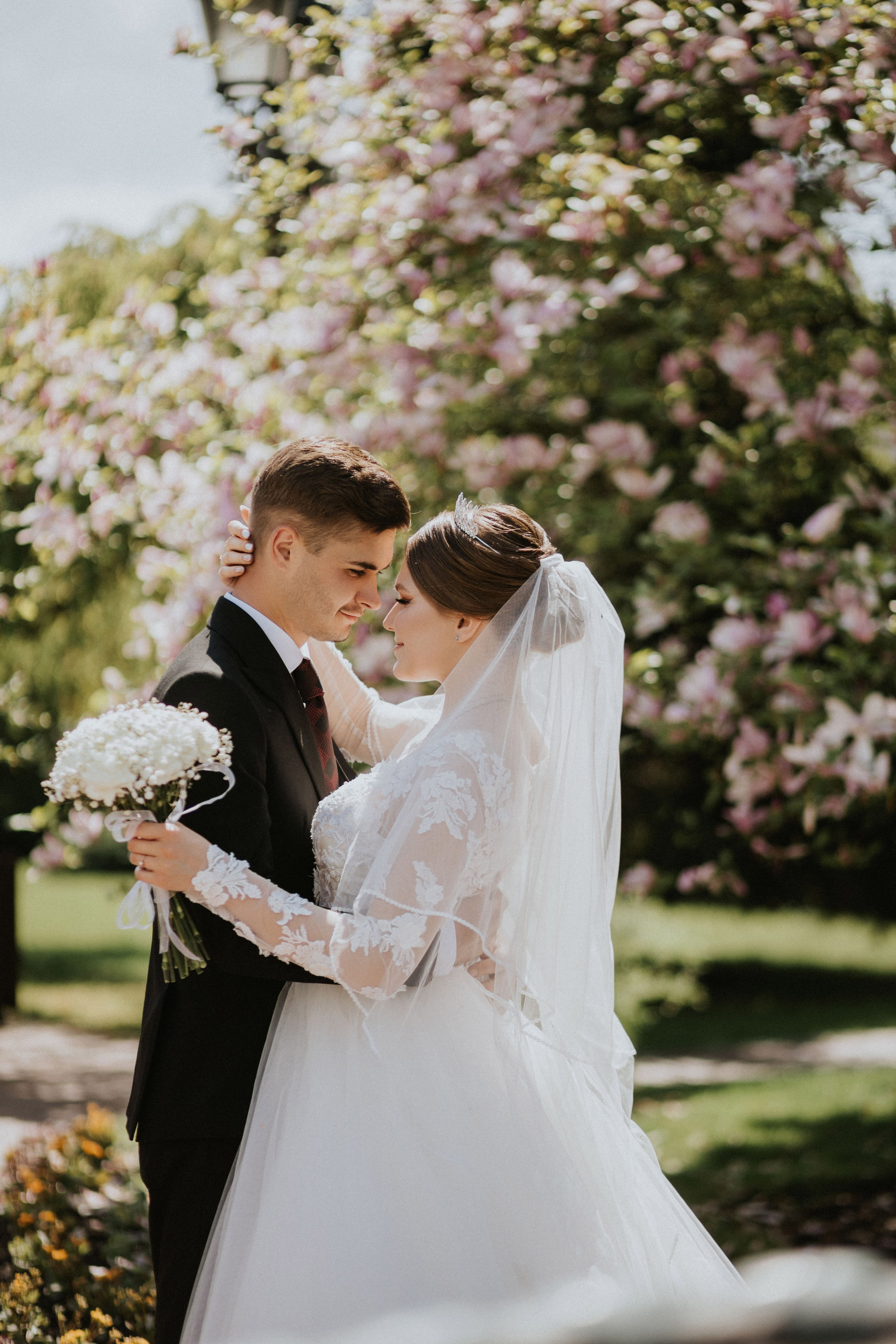 “Un romantico ritratto di matrimonio di una coppia innamorata, immortalato in un bellissimo giardino con fiori sbocciati sullo sfondo. La sposa, con in mano un bouquet di alito di bambino, guarda amorevolmente il suo sposo, mentre lui le tiene teneramente il viso. Entrambi sorridono, immersi in un momento gioioso e intimo”.