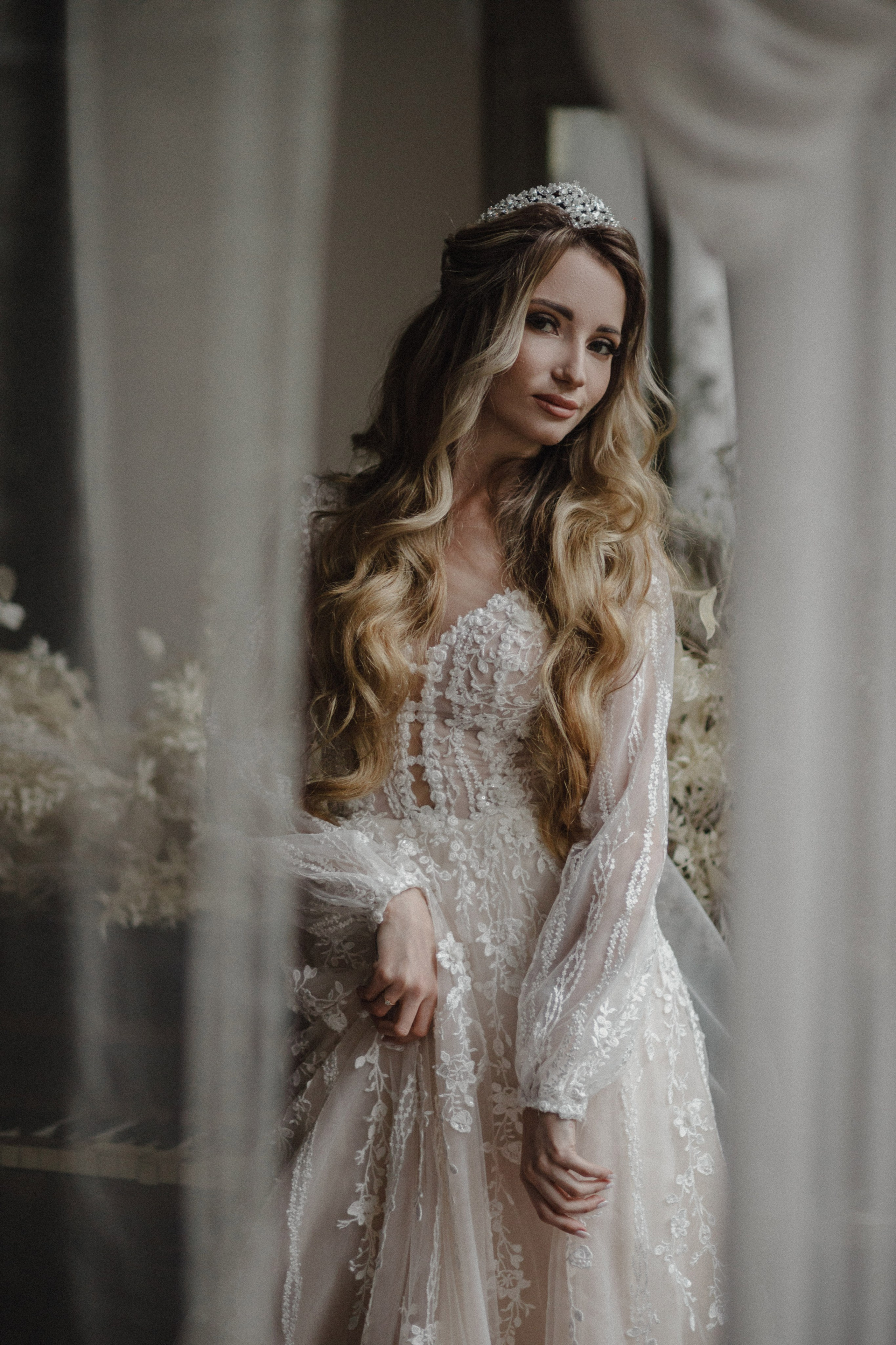 “Una sposa con lunghi capelli ondulati, che indossa un delicato diadema, si vede attraverso una tenda morbida e traslucida. Indossa un bellissimo abito da sposa in pizzo, con intricati dettagli floreali. L'illuminazione soffusa crea un'atmosfera serena ed eterea mentre lei guarda dolcemente la macchina fotografica”.