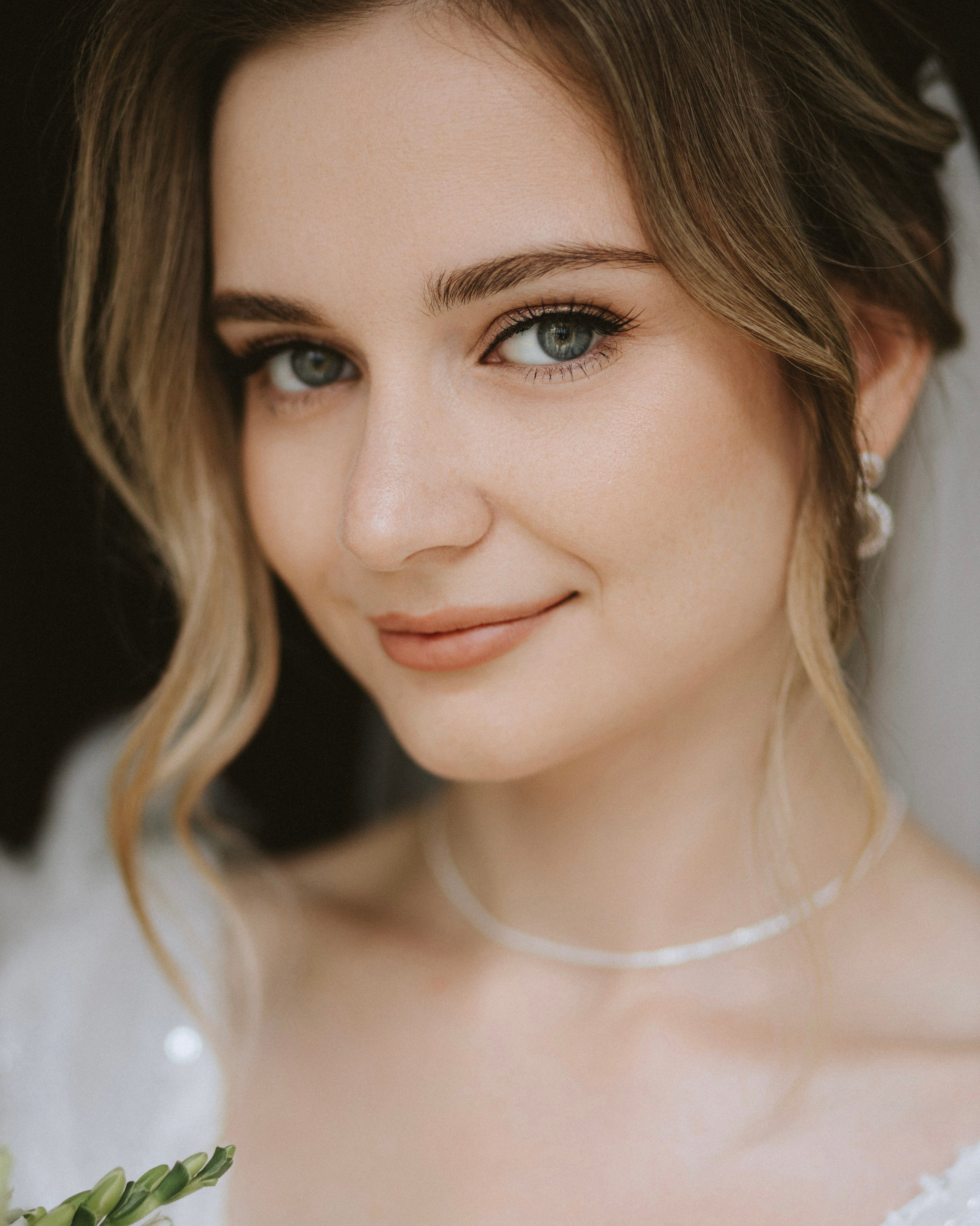 “The bride with soft makeup and elegant earrings, smiling against a dark background.”
