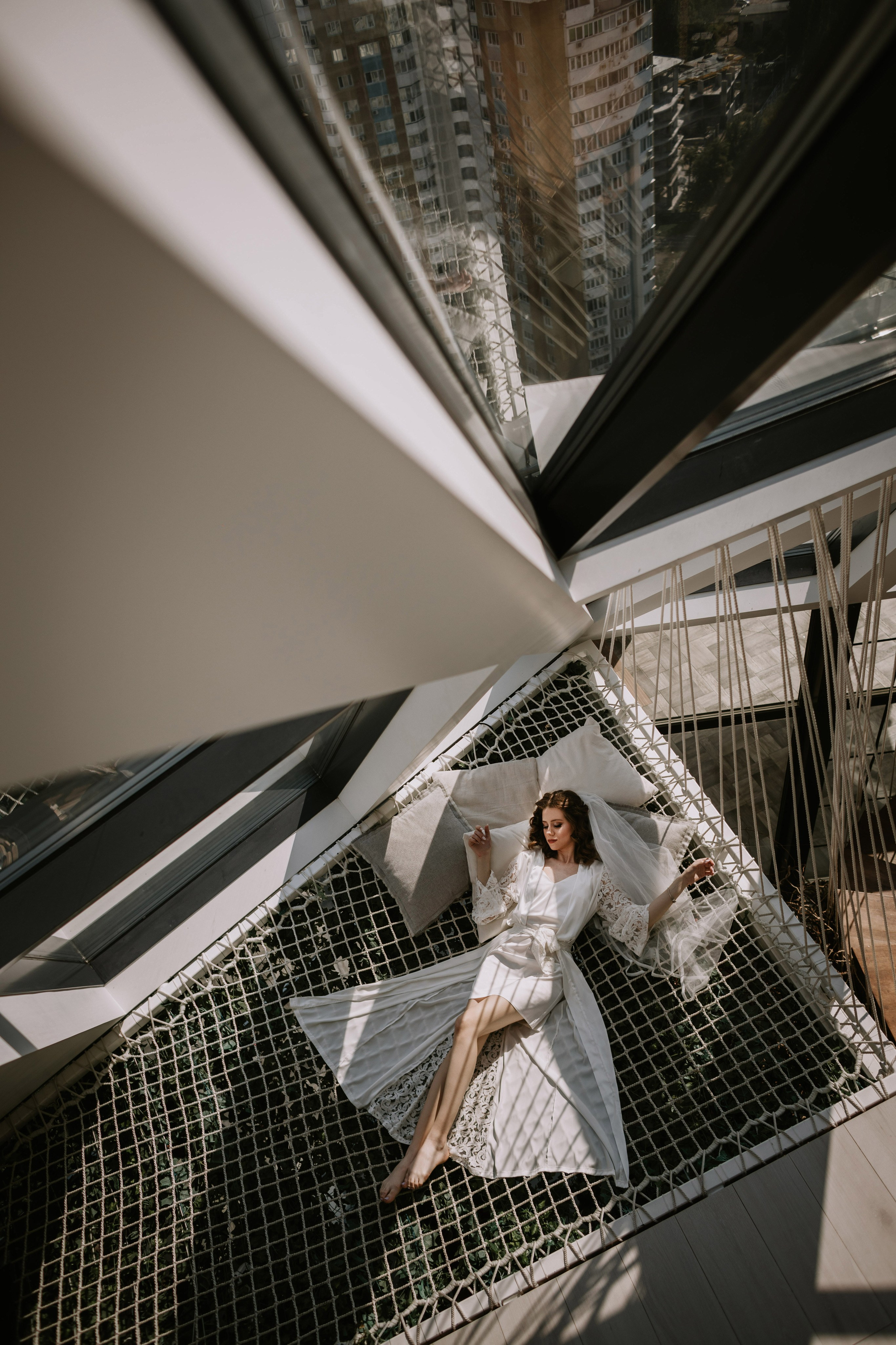 “Una sposa si adagia con grazia su un'amaca di rete all'interno di uno spazio moderno e aperto. La luce del sole filtra attraverso grandi finestre, proiettando motivi geometrici sul pavimento e circondandola di ombre morbide. Indossa una delicata vestaglia bianca con dettagli in pizzo e un velo, riposando con un braccio delicatamente appoggiato su un cuscino, mentre il paesaggio urbano si riflette nelle vetrate sopra di lei”.