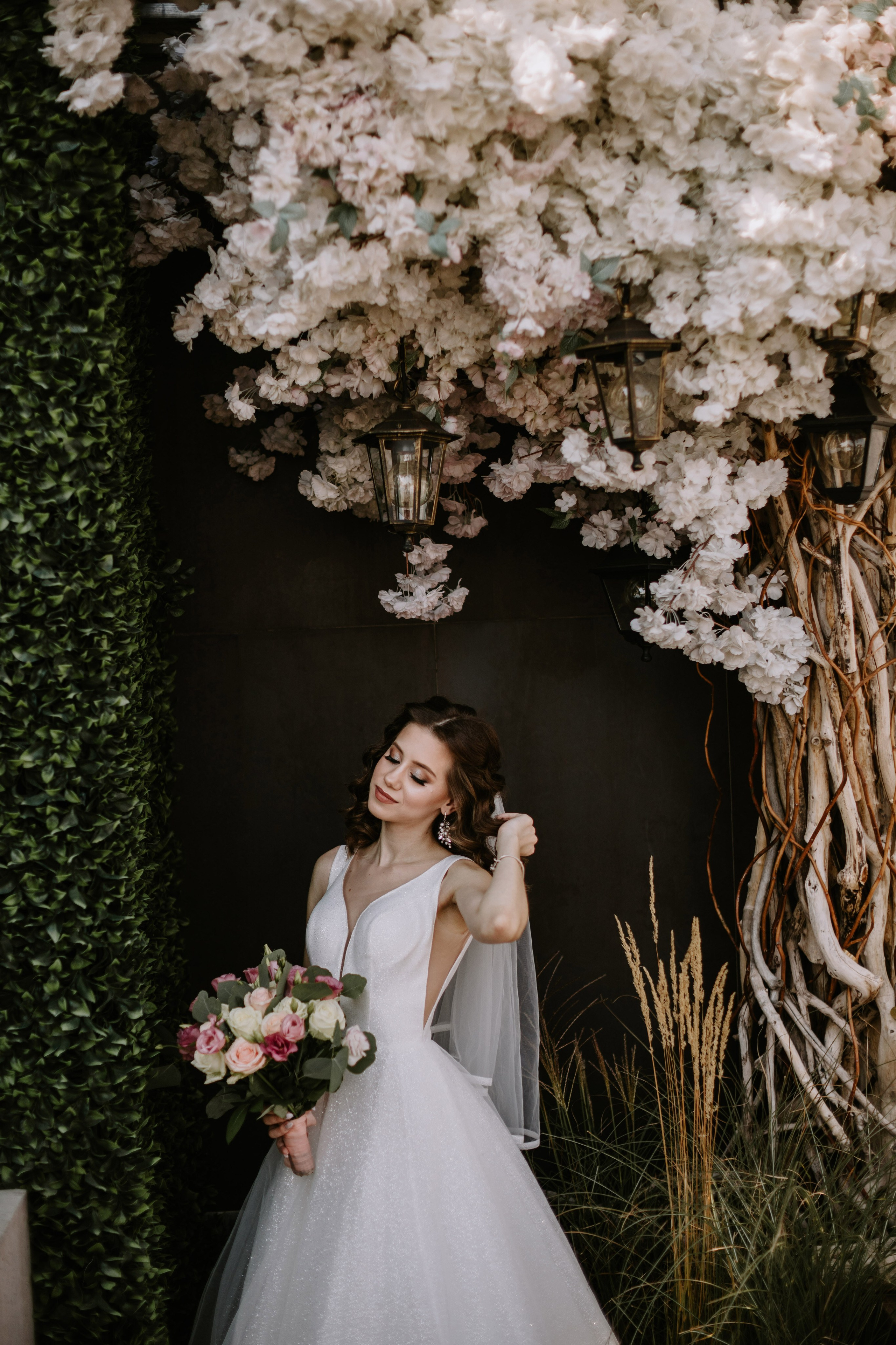 “Una sposa si erge con grazia sotto un arco floreale ornato da delicati fiori bianchi. Indossa un abito da sposa scintillante, senza maniche, con un profondo scollo a V e un velo fluente. I suoi capelli sono acconciati in morbide onde e regge un bouquet di fiori rosa, rosa e bianchi. Lo sfondo è ricco di vegetazione e le luci soffuse aggiungono un'atmosfera romantica alla scena”.