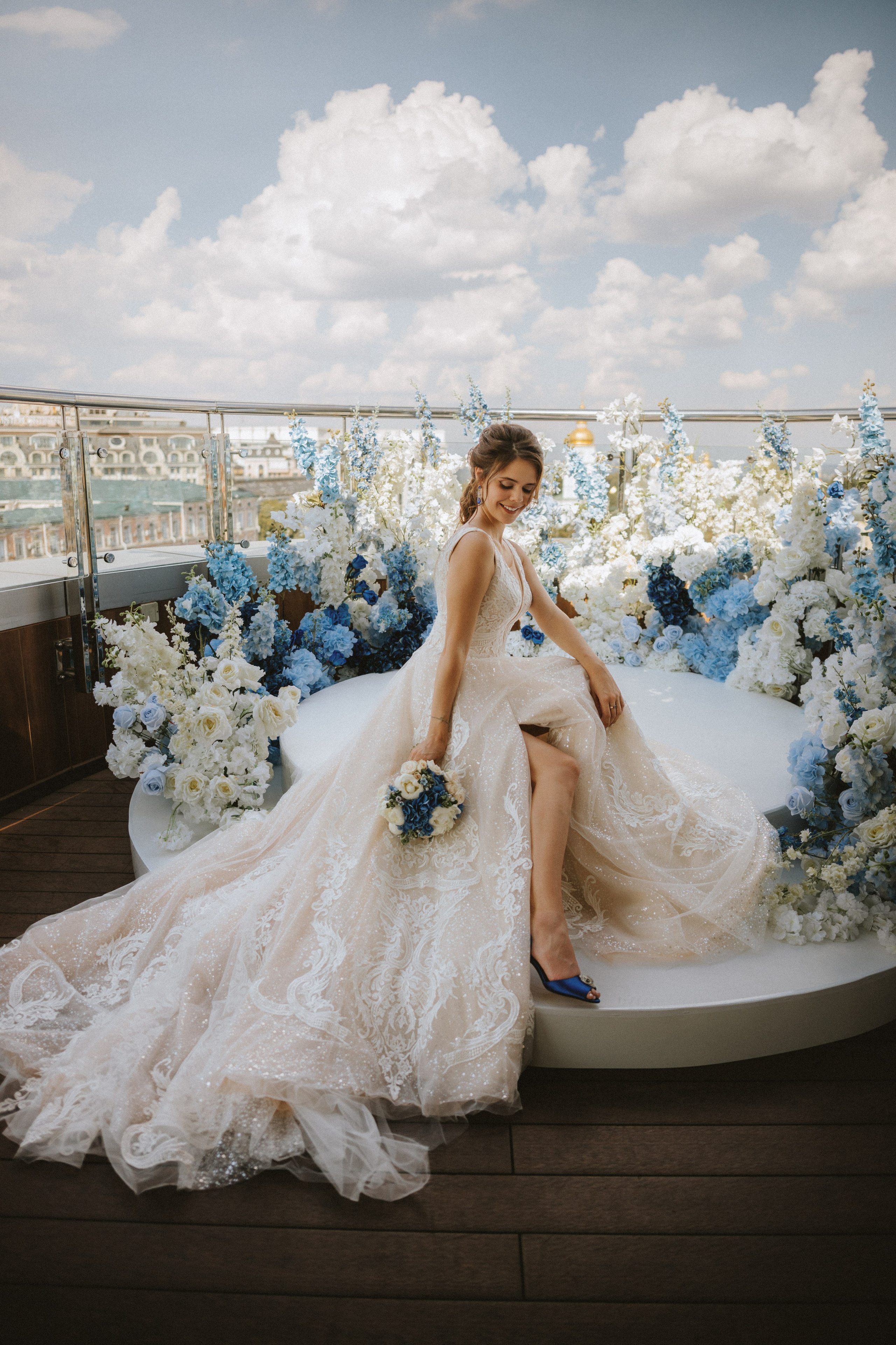 “Una sposa siede con grazia su una piattaforma circolare, circondata da una splendida serie di fiori bianchi e blu. Indossa un abito da sposa color avorio, scintillante, con intricati dettagli in pizzo e uno spacco all'altezza delle cosce. Il suo bouquet è in linea con il tema floreale, composto da fiori blu e bianchi. La sposa sorride e guarda in basso, con i capelli acconciati in un'elegante pettinatura. Sullo sfondo, il cielo è blu con nuvole soffici, che aggiungono un'atmosfera serena e romantica al momento”.