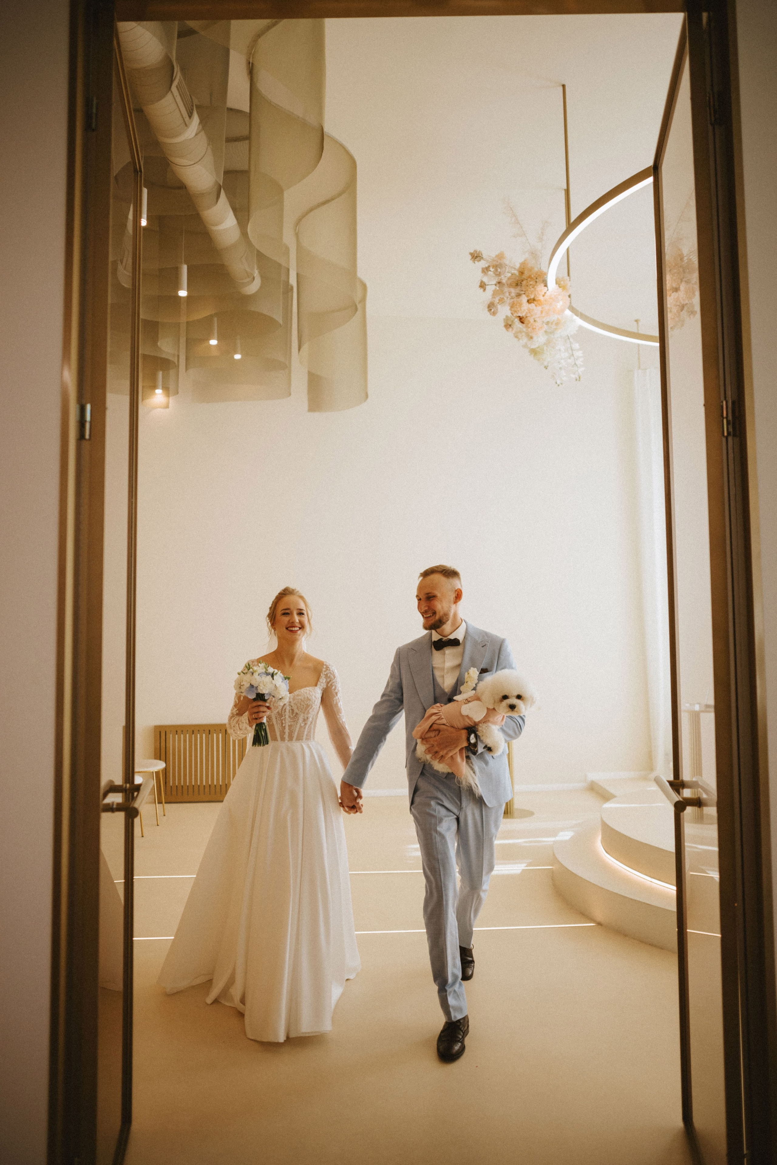 “Una coppia di sposi cammina mano nella mano in una stanza dal design moderno ed elegante, sorridendo con gioia. La sposa, in uno splendido abito di pizzo, tiene in mano un bouquet di fiori bianchi, mentre lo sposo, in abito azzurro e papillon, porta in braccio il loro soffice cane bianco. L'arredamento pulito e minimalista della sala, con luci soffuse e dettagli eleganti, esalta il momento celebrativo”.