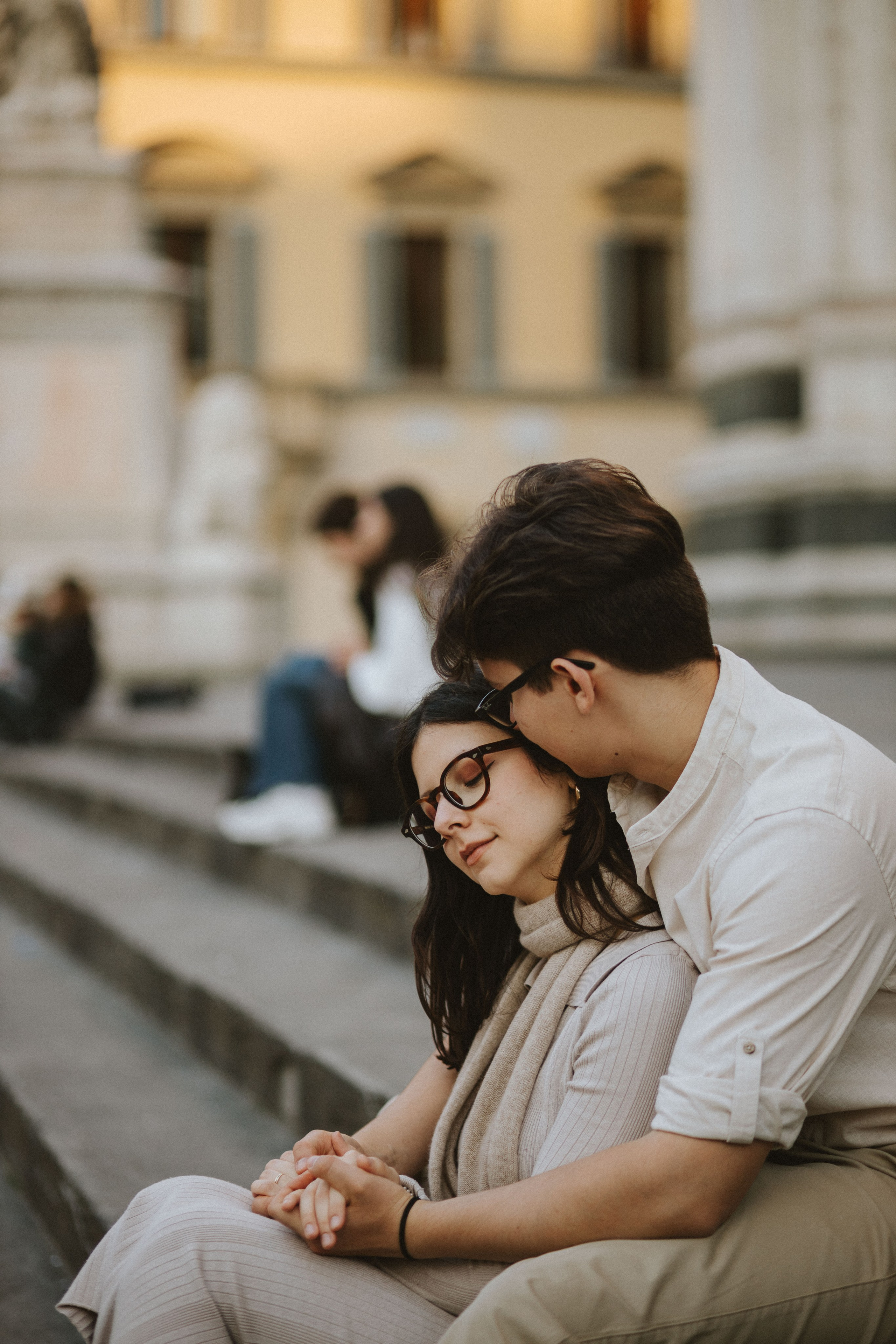“Una coppia seduta su gradini di pietra in un luogo panoramico, con l'uomo che appoggia delicatamente la testa sulla spalla della donna mentre si tengono per mano. La donna ha gli occhi chiusi ed entrambi sorridono dolcemente. Sullo sfondo, persone che camminano e siedono nella piazza”.