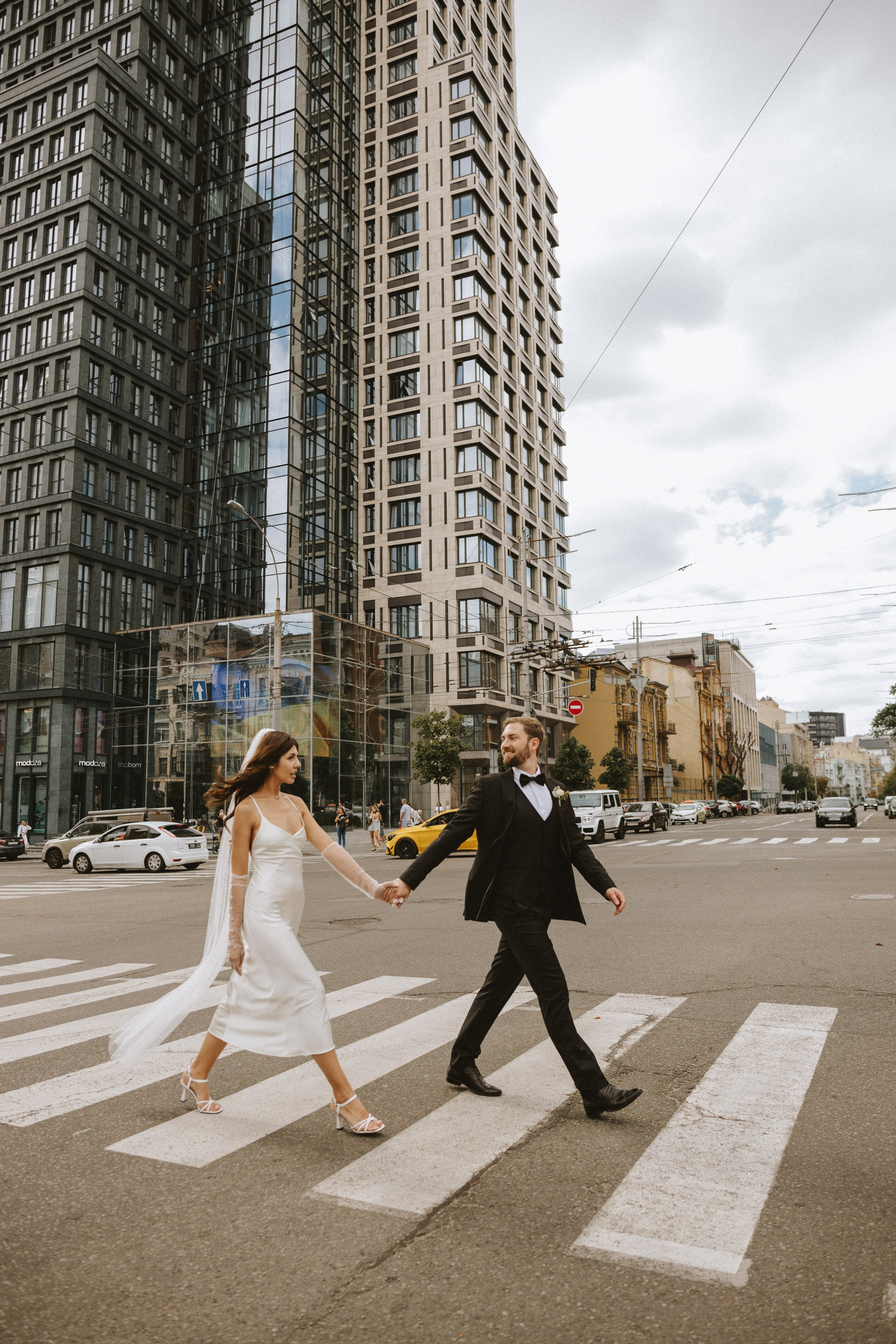 “Una sposa e uno sposo camminano mano nella mano su un passaggio pedonale in un contesto urbano, circondato da alti edifici moderni. La sposa indossa un elegante abito bianco con un lungo velo, mentre lo sposo sfoggia un classico smoking nero. Entrambi sorridono, godendosi un momento di gioia nell'atmosfera frenetica della città”.