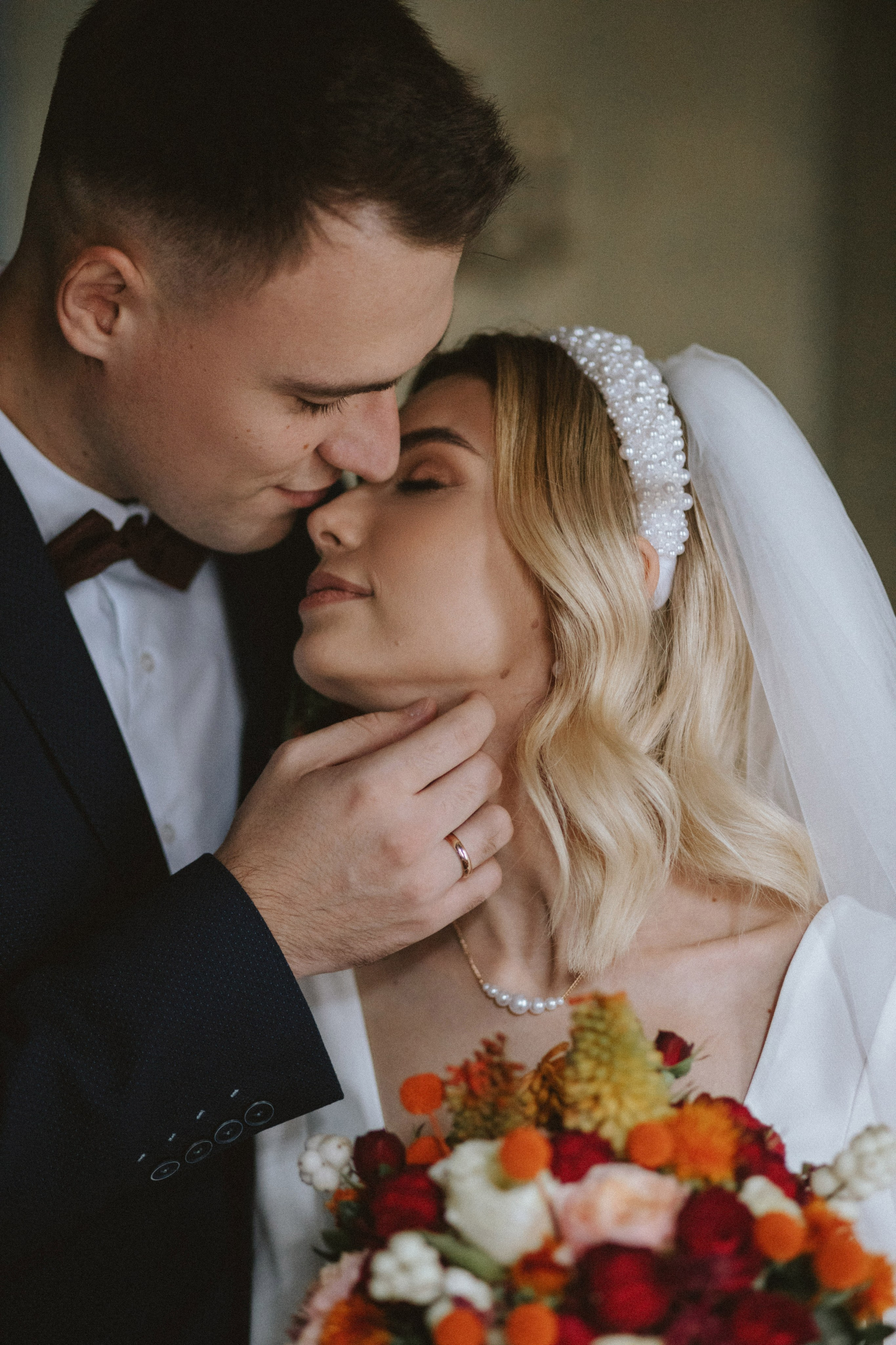 “Un primo piano di una sposa e di uno sposo che condividono un momento di intimità, con i loro volti che quasi si toccano. Lo sposo tiene delicatamente il mento della sposa, mentre lei chiude gli occhi, irradiando amore e tenerezza. La sposa indossa un bellissimo cerchietto di perle e il suo bouquet di fiori vivaci aggiunge un tocco di colore a questo momento caldo e romantico”.