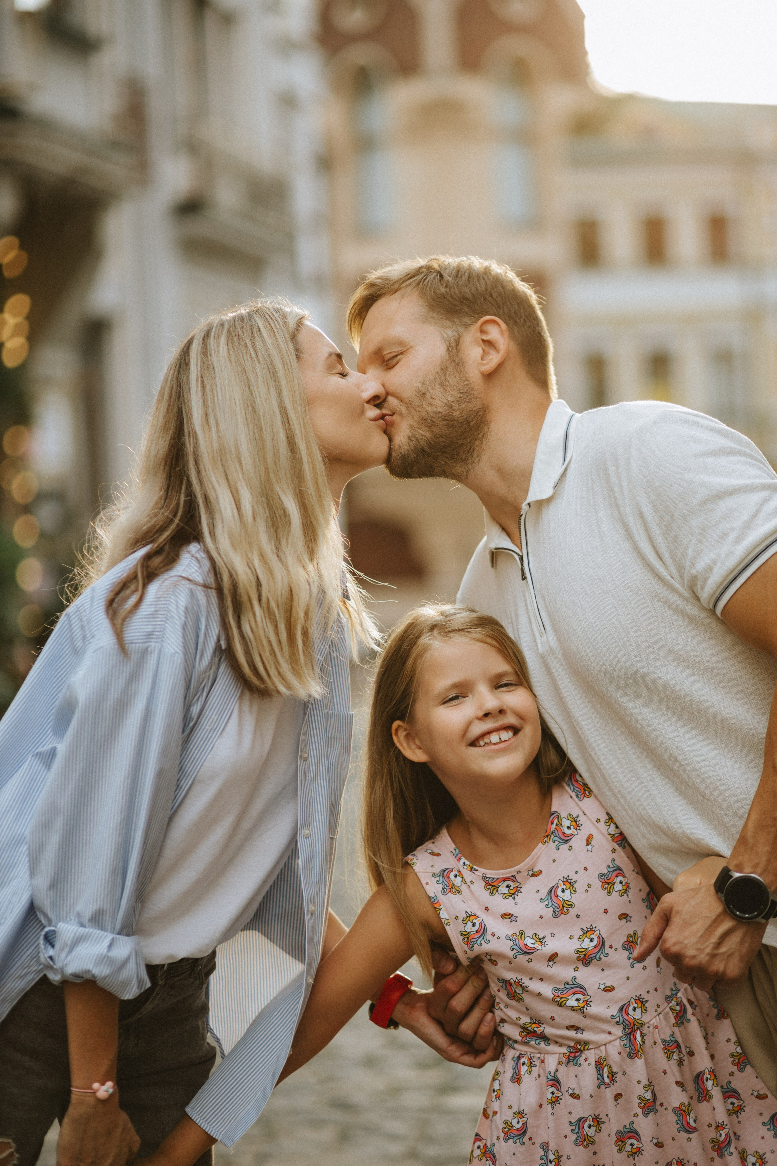 “Una famiglia felice in piedi insieme in una strada illuminata dal sole. Il padre e la madre si scambiano un tenero bacio, con le teste protese l'una verso l'altra in un gesto d'amore. Di fronte a loro, la figlia piccola, che indossa un giocoso vestito rosa con stampe di unicorni, sorride gioiosa all'obiettivo. La scena cattura il calore e l'affetto, con lo sfondo che mostra una morbida sfocatura di edifici urbani, evidenziando lo stretto legame tra i membri della famiglia”.