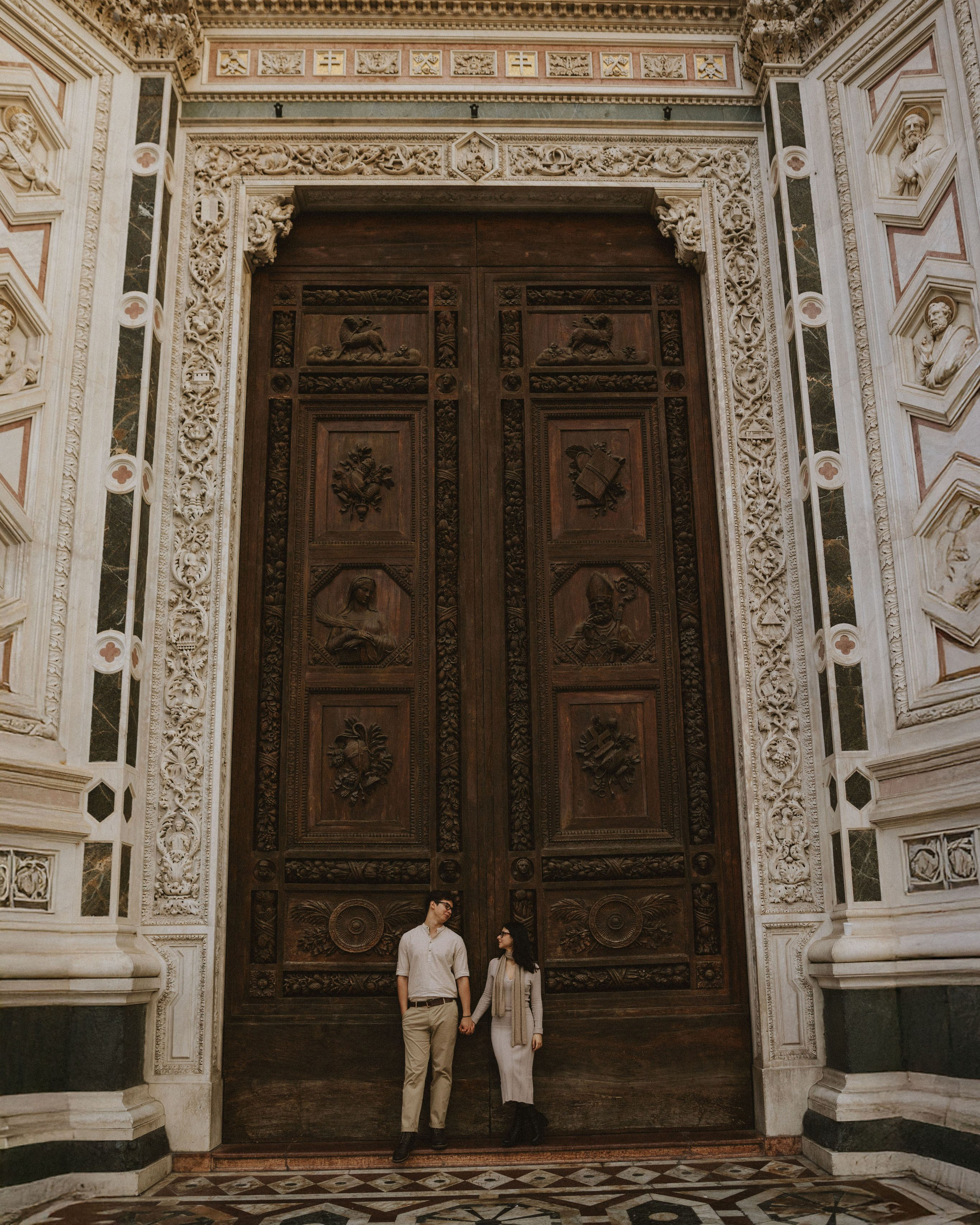 “A couple standing by large wooden doors with stone architecture and intricate ornamentation, holding hands.”