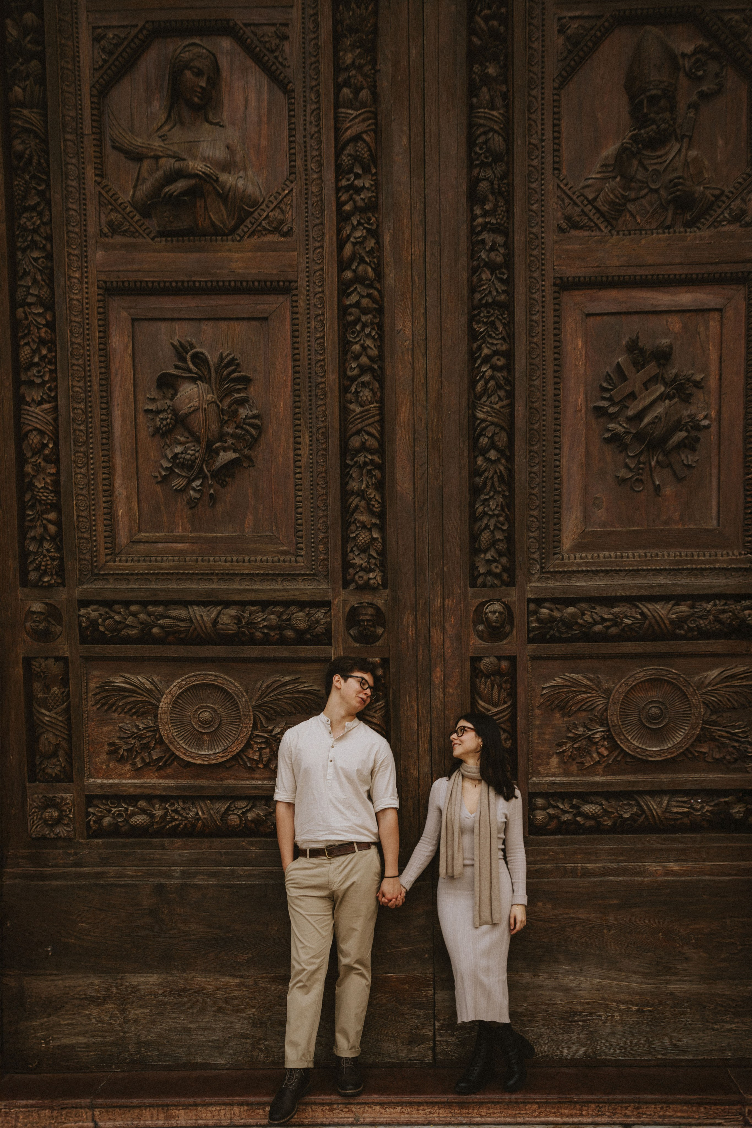 “Una coppia in piedi davanti a una grande porta di legno intricatamente intagliata, che si tiene per mano e si guarda sorridendo”.