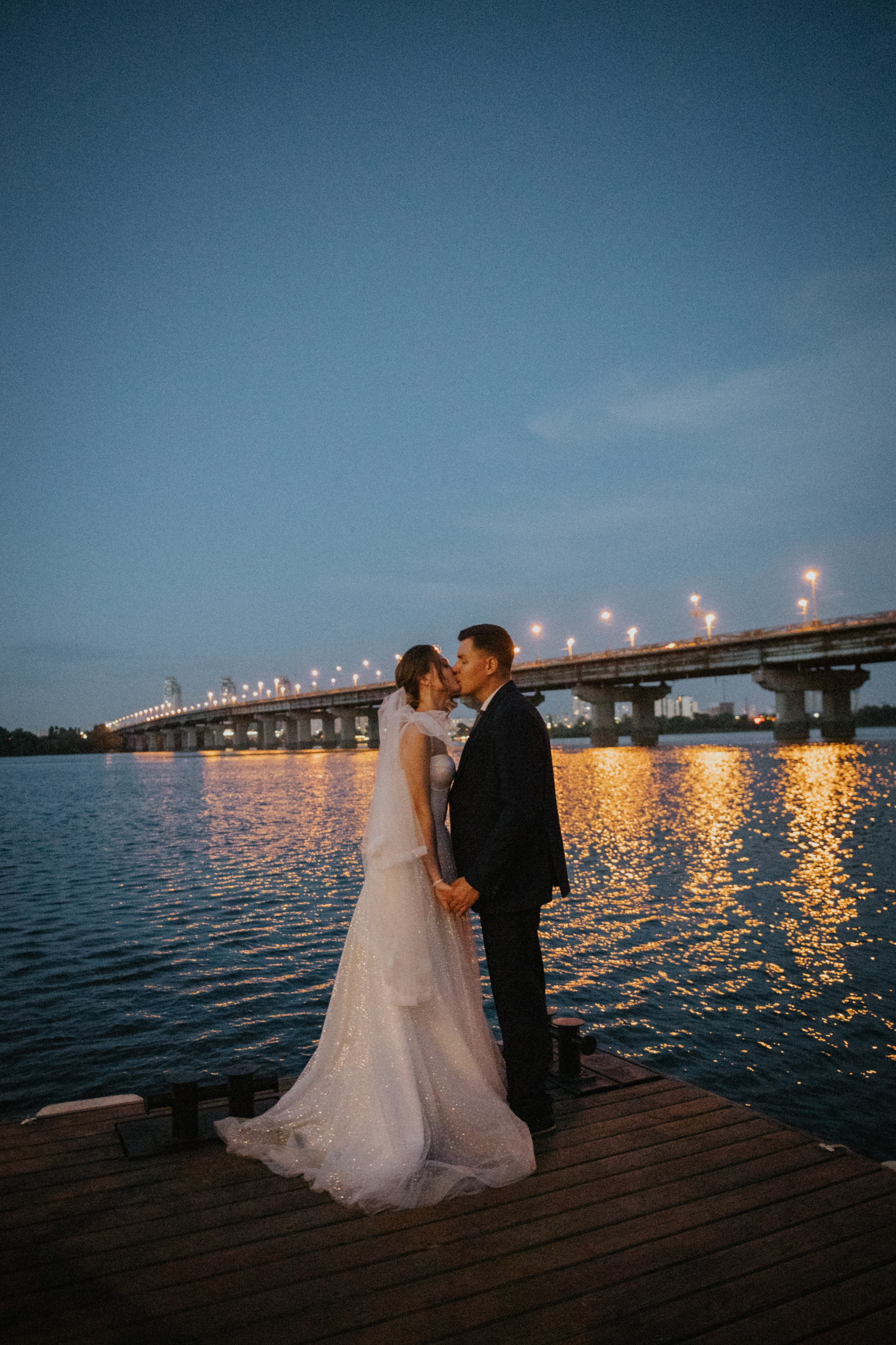 “Una coppia si trova su un molo in riva al fiume, tenendosi per mano e scambiandosi un bacio sotto il cielo notturno. Le luci scintillanti di un ponte vicino si riflettono sull'acqua, creando un'atmosfera romantica e serena. La sposa, vestita con un abito da sposa scintillante, è avvolta da una luce soffusa, mentre lo sposo, in abito scuro, abbraccia il momento con la sua sposa”.