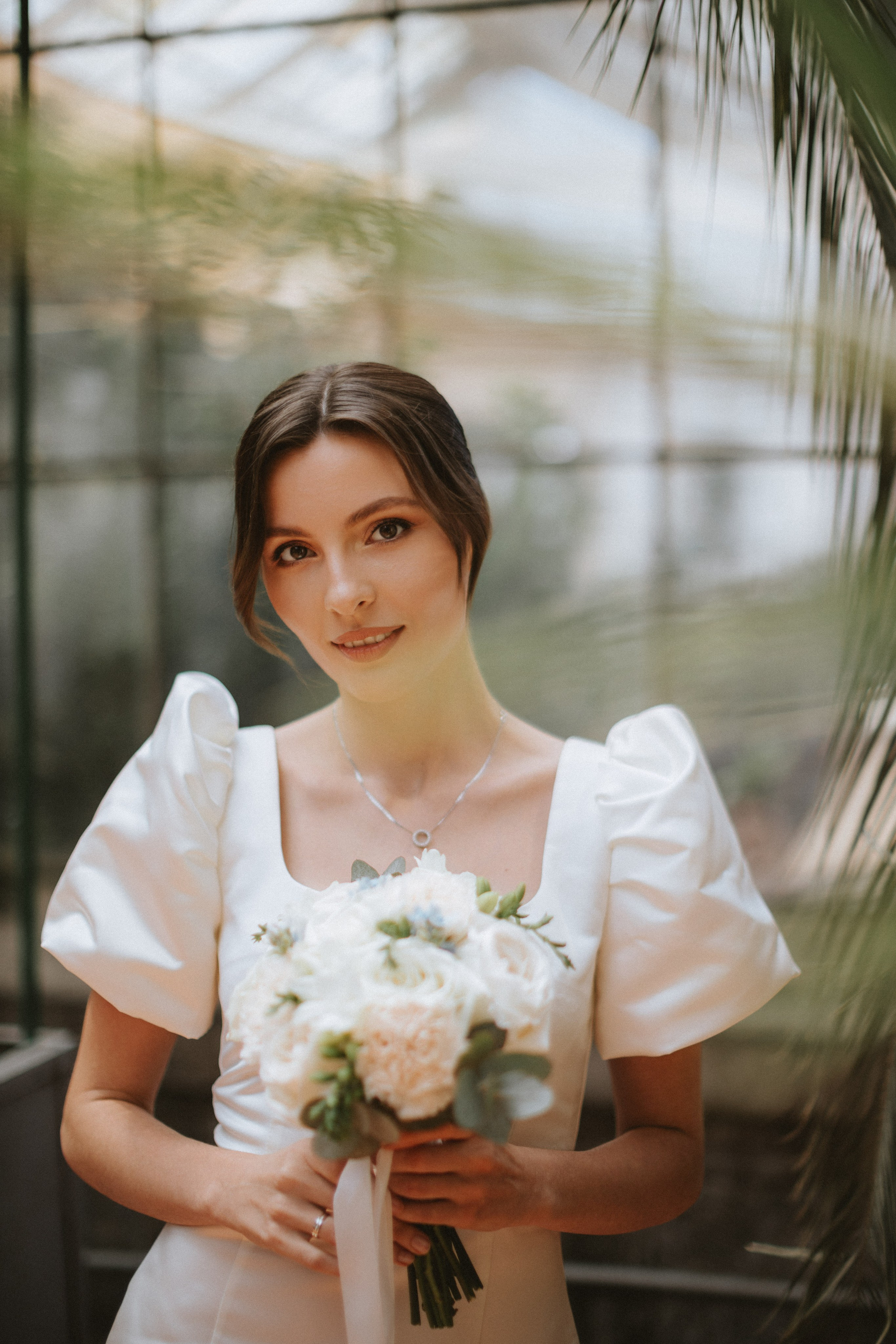 “Una sposa dai corti capelli castani, che indossa un abito bianco con maniche a sbuffo, sorride dolcemente tenendo in mano un bouquet di fiori bianchi e rosa chiaro. Si trova in una serra circondata da piante, con un'espressione dolce e serena”.