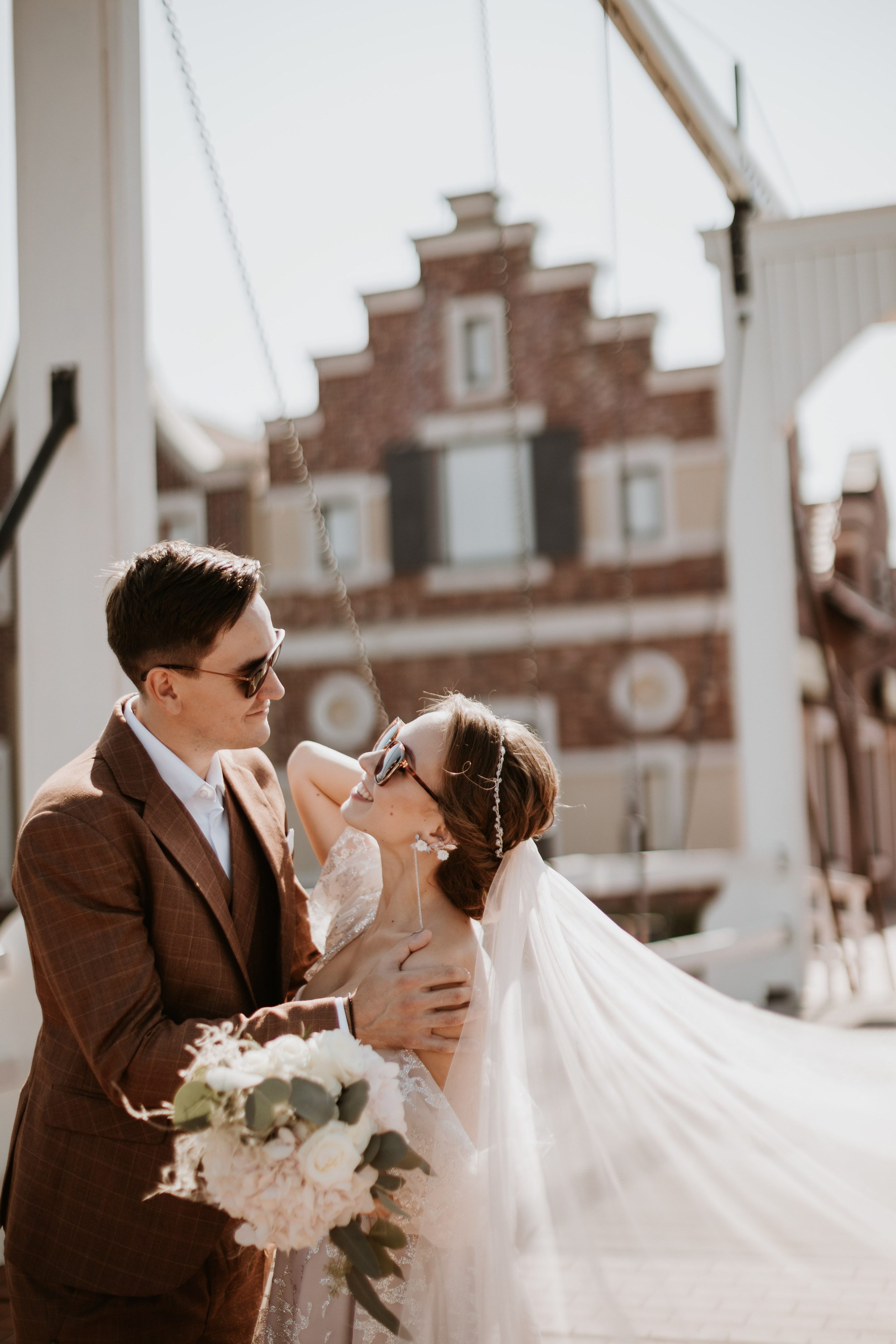 “Una coppia felice si abbraccia in un momento giocoso e gioioso all'aperto. La sposa, in abito da sposa e velo, tiene in mano un bouquet di fiori bianchi, mentre lo sposo, in abito marrone, le sorride. Entrambi indossano occhiali da sole, aggiungendo un tocco casual e moderno alla classica scena del matrimonio. Sono in piedi davanti a un incantevole edificio, con la luce soffusa del sole che mette in risalto i loro sorrisi e il velo fluente, creando un'atmosfera romantica e spensierata”.