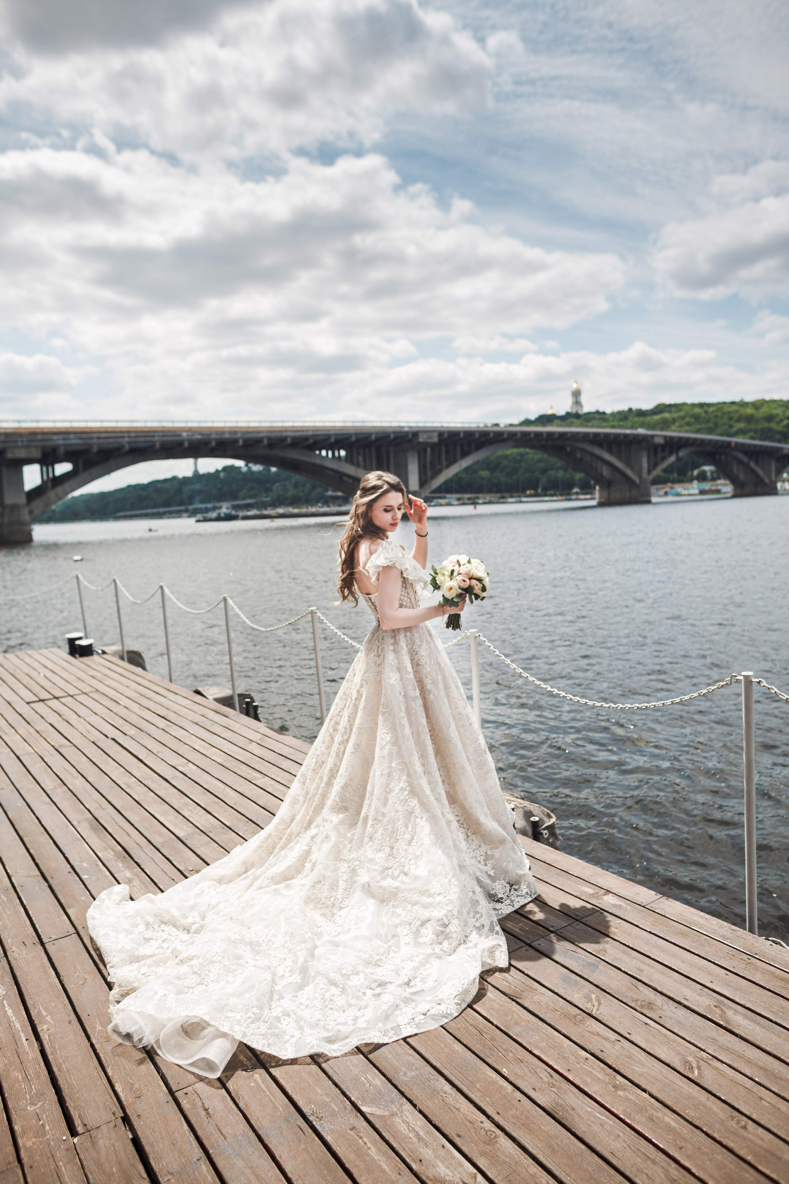 “Una bella sposa si trova su un molo di legno lungo un fiume, con un ponte sullo sfondo. Indossa un intricato abito da sposa in pizzo con un lungo strascico e tiene in mano un bouquet di rose rosa pallido. Il vento solleva delicatamente il suo velo, aggiungendo un senso di movimento alla scena serena e romantica. Il cielo è parzialmente nuvoloso e le acque calme riflettono l'ambiente pittoresco”.