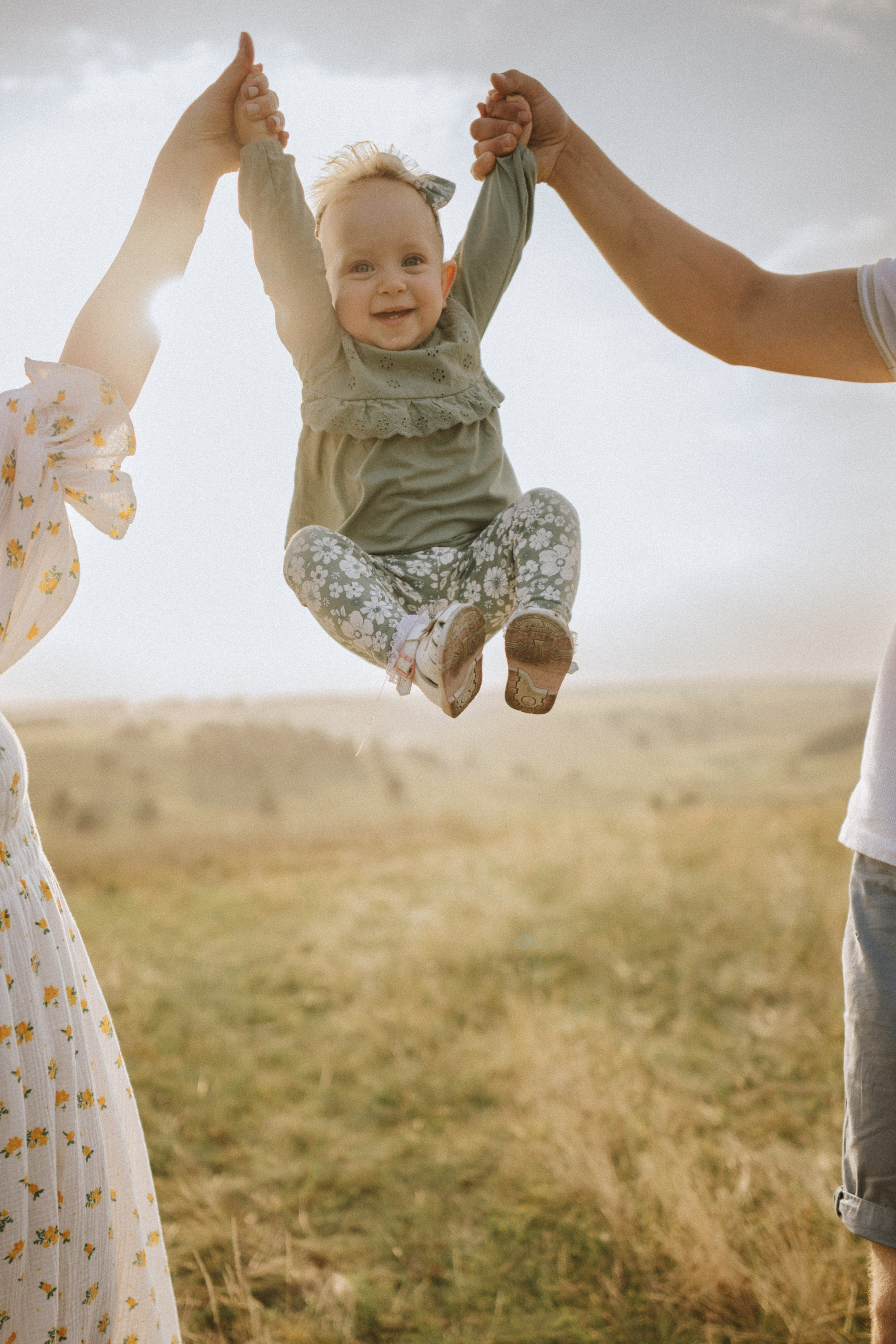 “Una bambina gioiosa con un top verde e pantaloni a fiori, sollevata dai genitori in una giornata di sole in un campo erboso. La bambina sorride, con le braccia alzate, mentre i genitori le tengono le mani, con il sole che splende sullo sfondo”.