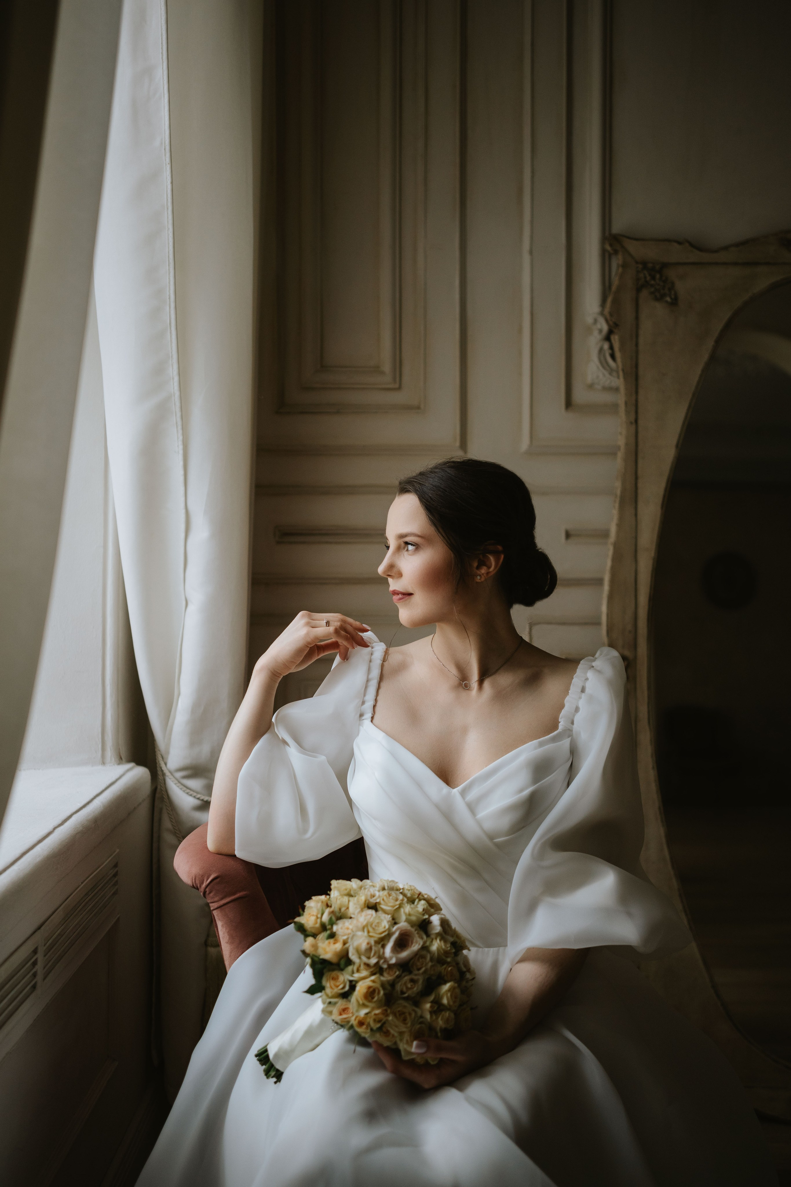 “Fotografo di matrimonio che immortala una bellissima sposa in un elegante abito da sposa, seduta accanto a una finestra e con in mano un bouquet di rose gialle. Il momento sereno è ambientato in un interno elegante, perfetto per un matrimonio in Toscana, Italia”.