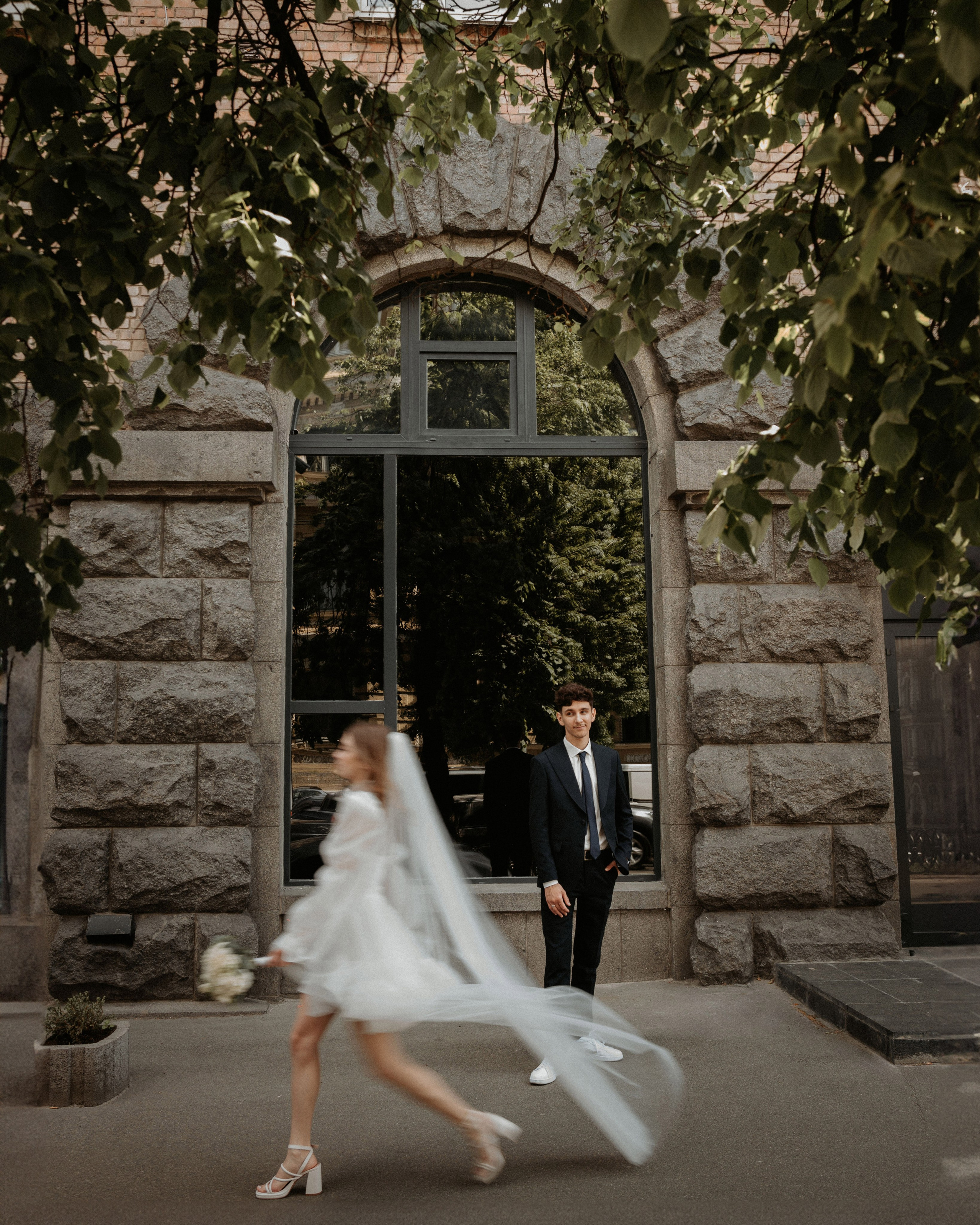 “Uno scatto di matrimonio giocoso ed energico con una sposa che indossa un abito bianco corto e scarpe da ginnastica, con il velo che scorre mentre passa davanti allo sposo, che rimane fermo e le sorride. Lo sfondo mostra un vecchio edificio in mattoni con grandi finestre, incorniciato dalle foglie verdi di un albero, che aggiungono un'atmosfera vibrante e dinamica alla scena”.