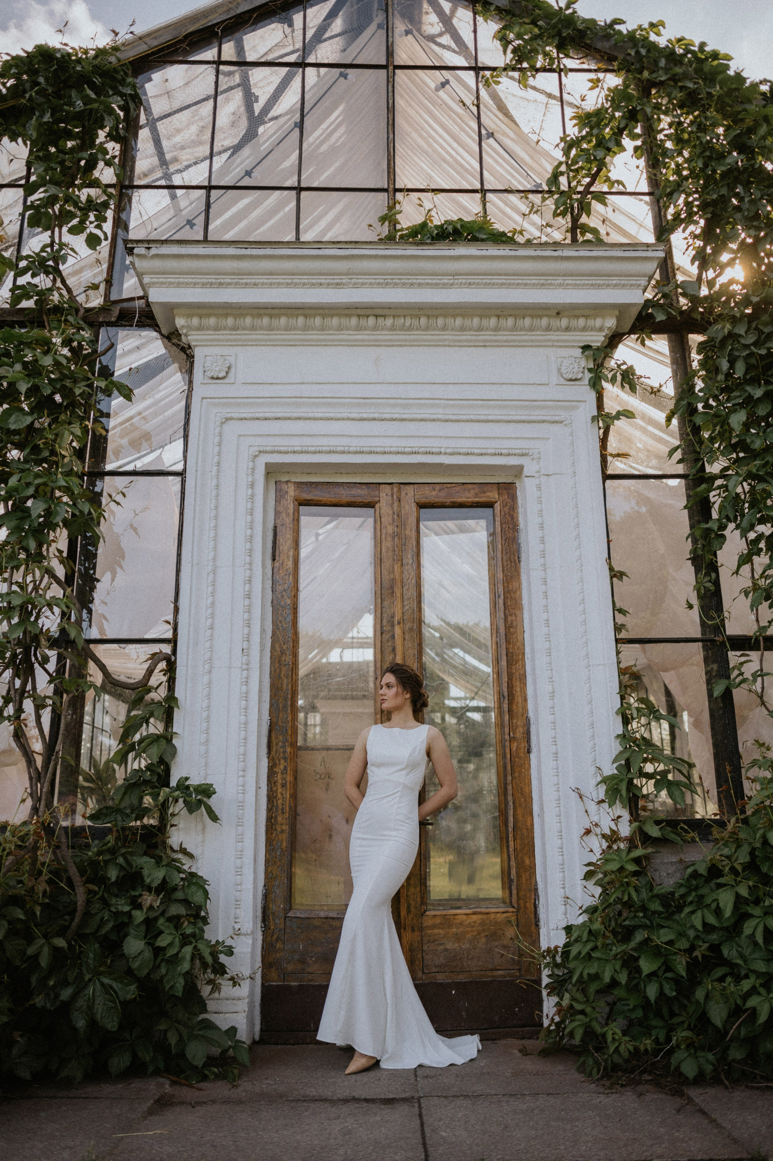 “Una donna con un abito da sposa bianco e aderente sta elegantemente davanti a una porta di legno rustica incorniciata dal verde e da una struttura a serra di vetro. La donna appoggia la mano sul telaio della porta e guarda di lato, con i capelli acconciati in morbide onde. Il sole illumina delicatamente la scena, creando un'atmosfera calda e serena”.