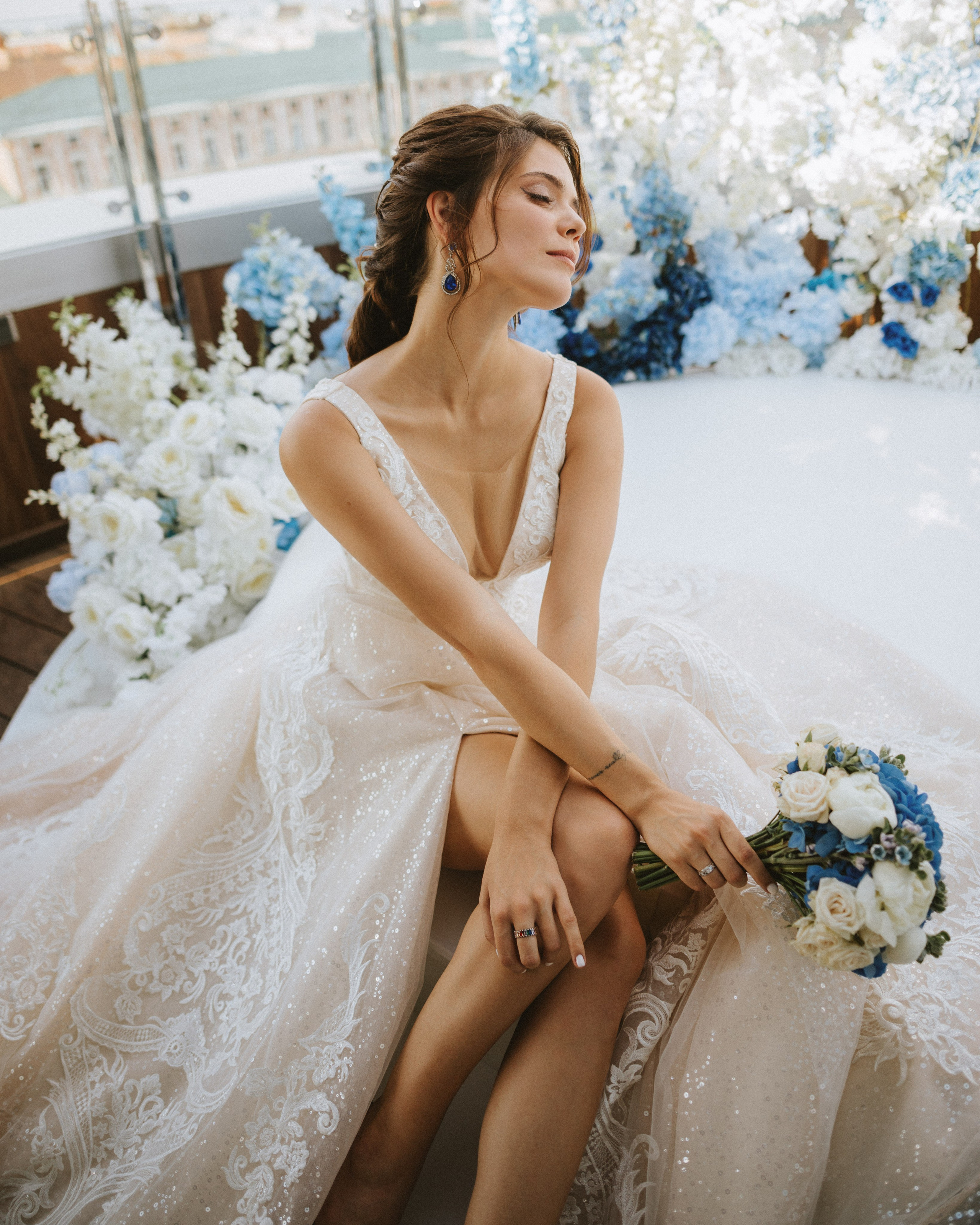 “The bride in an exquisite dress with a bouquet of white and blue flowers, sitting among luxurious flowers at a wedding in Sardinia or the Amalfi Coast.”
