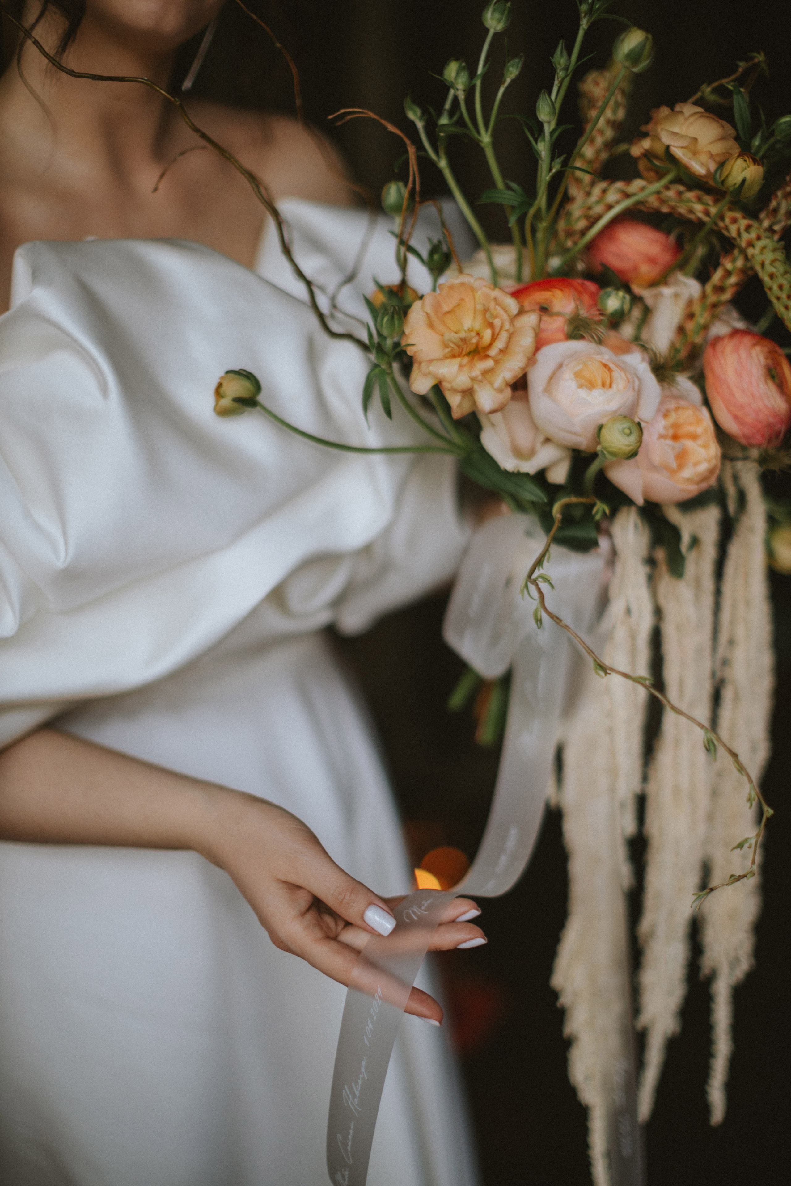 “Primo piano di una sposa che tiene in mano un bellissimo bouquet di fiori rosa e pesca, tra cui ranuncoli, con delicati nastri che scendono dai gambi. La mano della sposa tiene delicatamente i nastri, con morbide unghie bianche che completano il suo elegante abito di raso”.