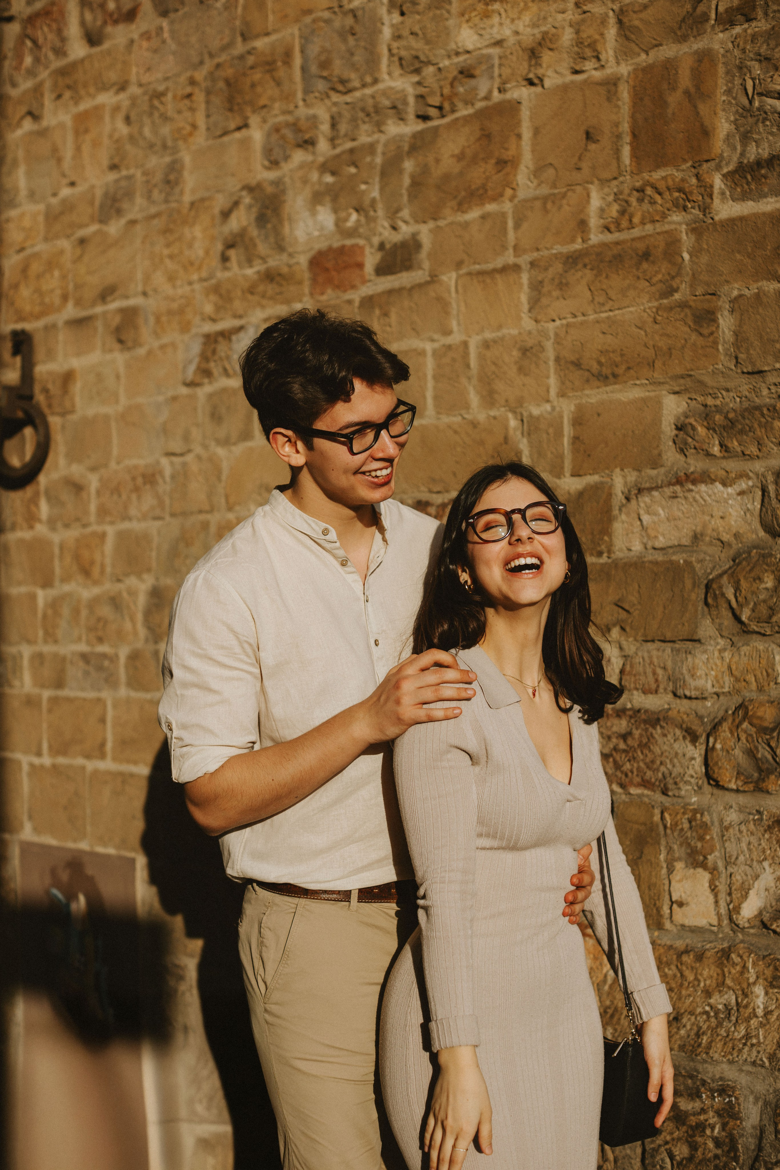 “Una coppia che sorride e ride insieme, in piedi davanti a un muro di mattoni alla luce del sole”.