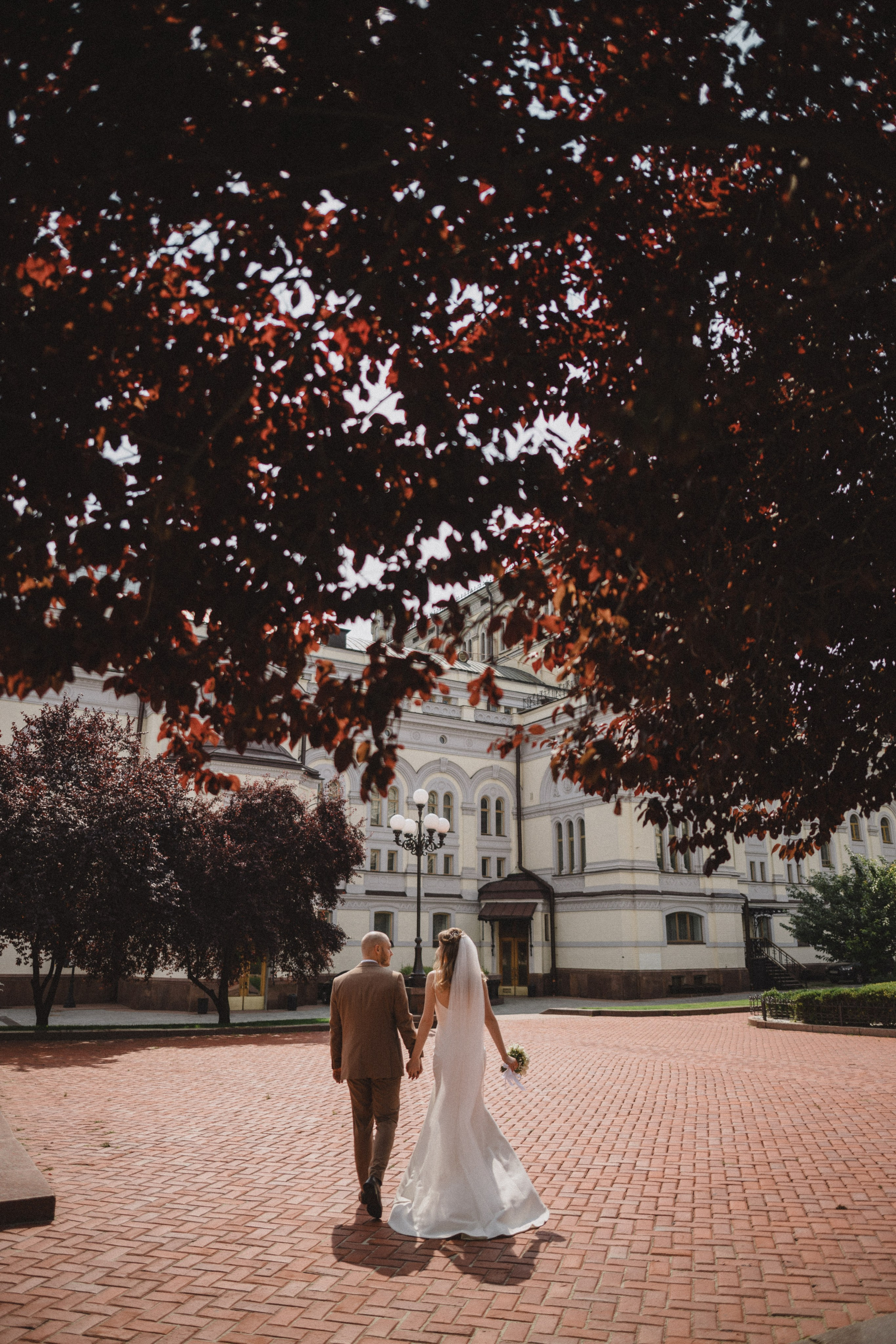 “Una coppia cammina mano nella mano, con la sposa che indossa un fluente abito bianco e un velo, mentre lo sposo indossa un abito marrone chiaro. Passeggiano sotto un albero dalle foglie bordeaux intenso, sullo sfondo di un edificio storico. La scena è illuminata da una luce solare soffusa, che crea un'atmosfera calda e romantica”.
