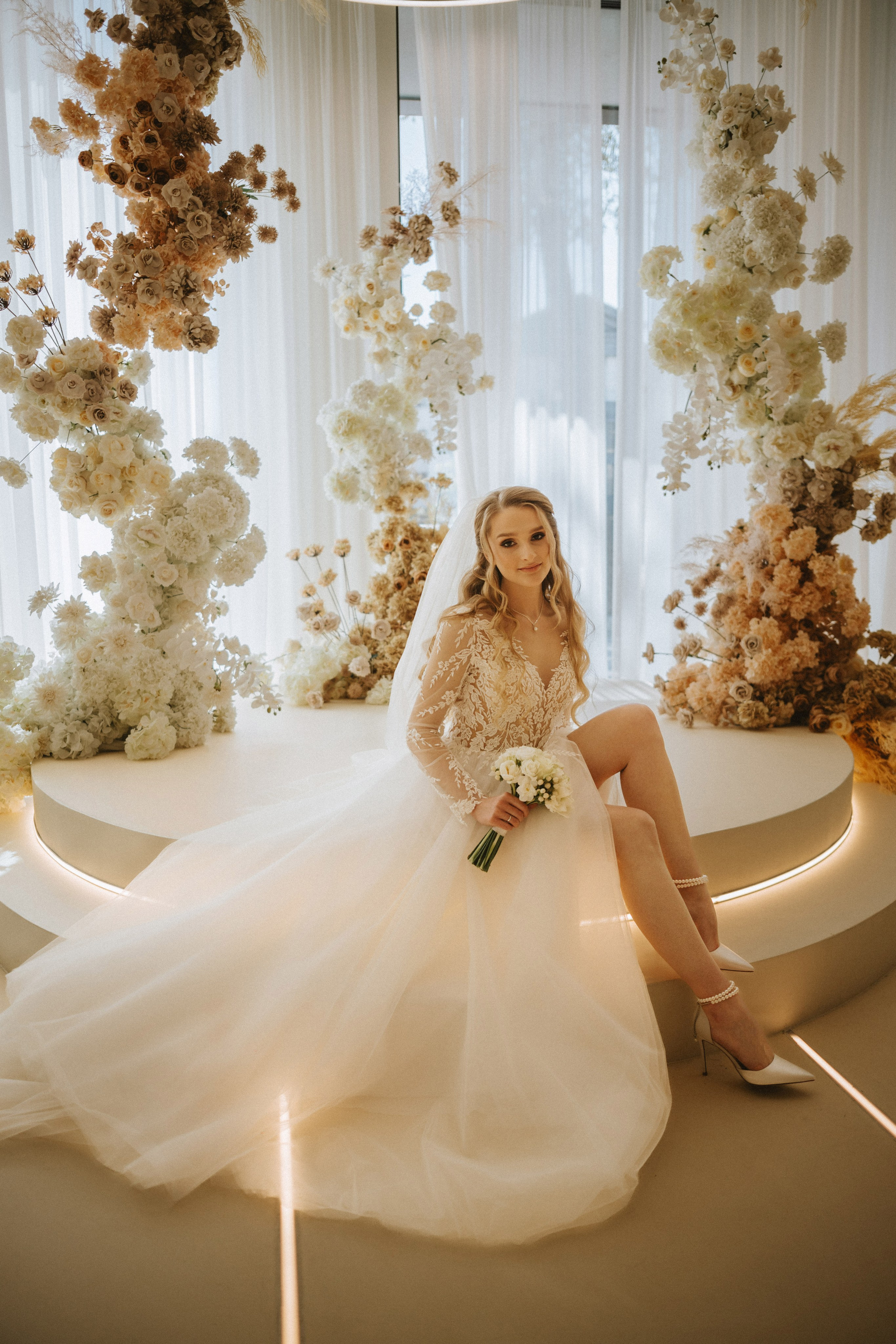 “La sposa siede con grazia in mezzo a una splendida esposizione di fiori bianchi e crema, tenendo tra le mani un delicato bouquet. La luce soffusa mette in risalto il suo elegante abito da sposa con intricati dettagli in pizzo, creando un'atmosfera serena ed elegante perfetta per un matrimonio sofisticato.”